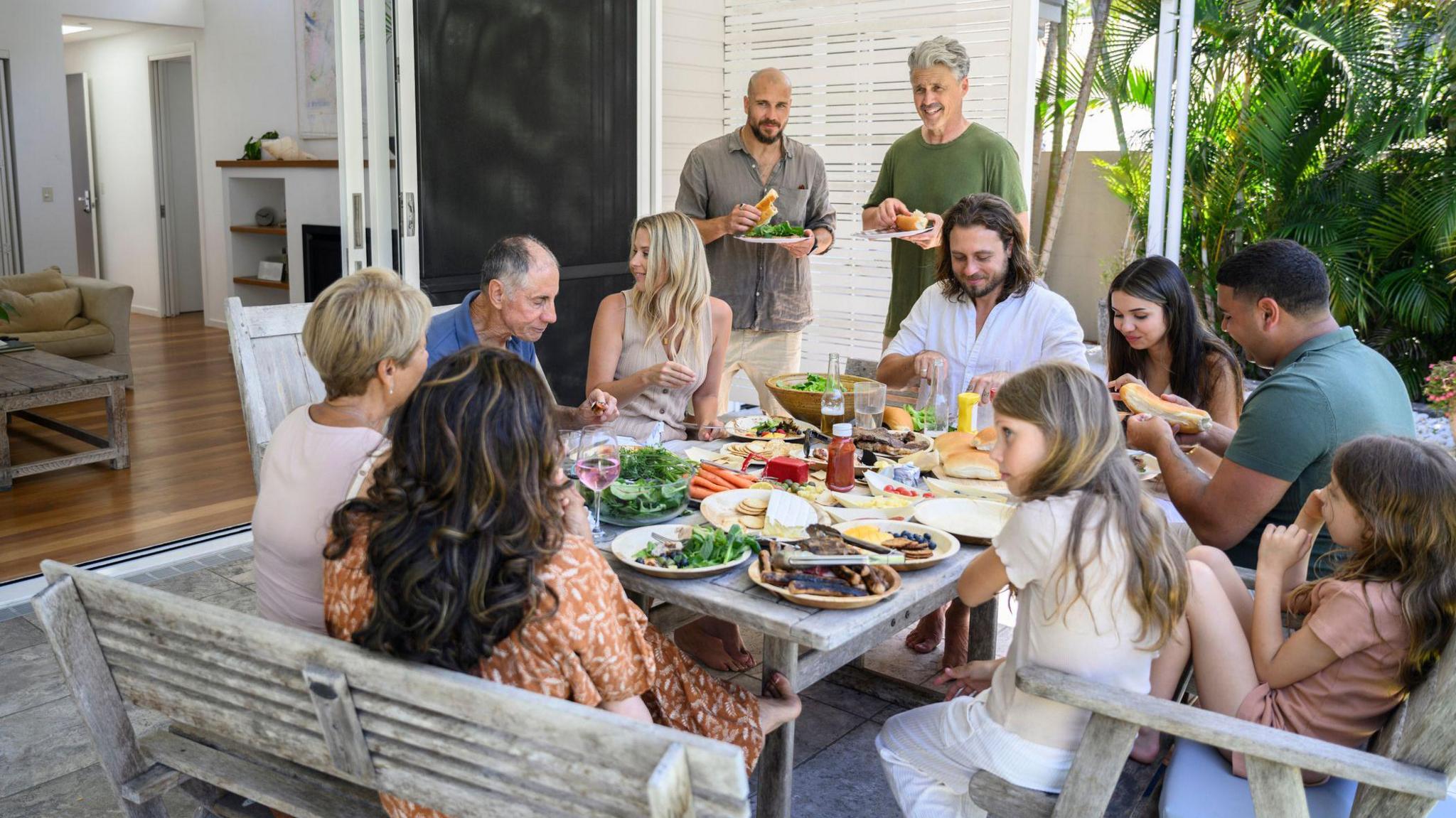 9 people sit and eat a BBQ on around the table. Two people are standing and eating burgers in the background