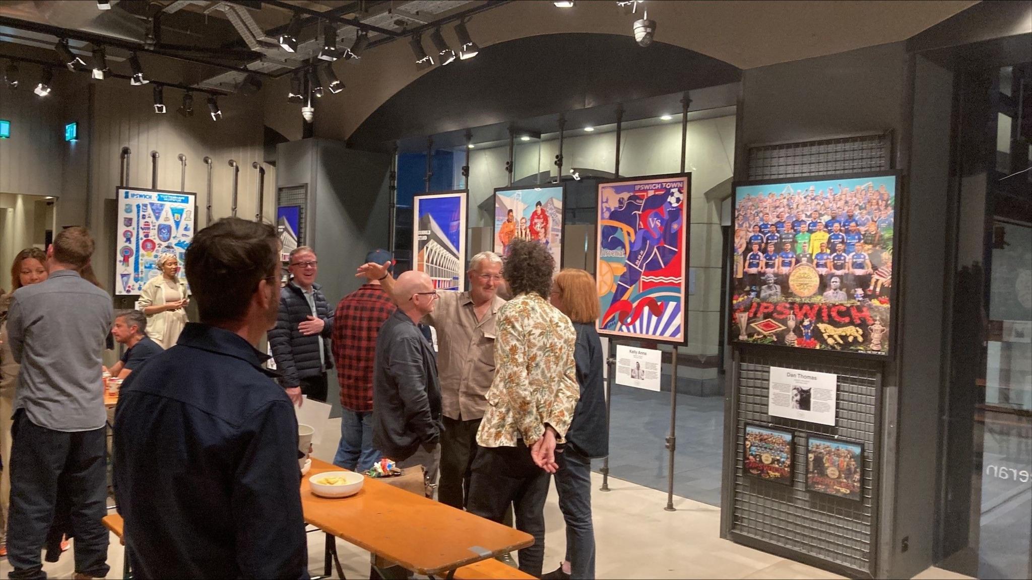 A group of people standing in a room that is full of Ipswich Town posters, in frames around the room.