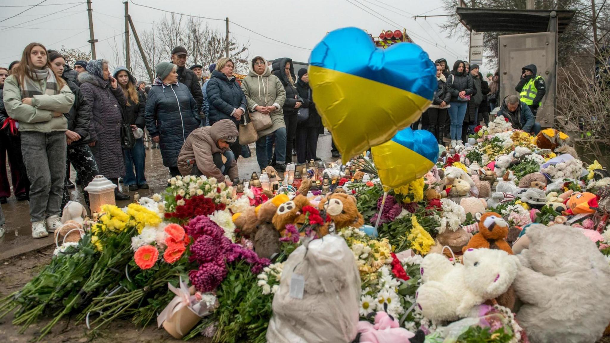 Dozens of people, wearing hats and large jackets, gather around a bed of flowers laid on the ground as a memorial to victims of a Russian strike. Among the flowers, there are cuddly toys and balloons with the Ukrainian flag in the shape of a heart. 