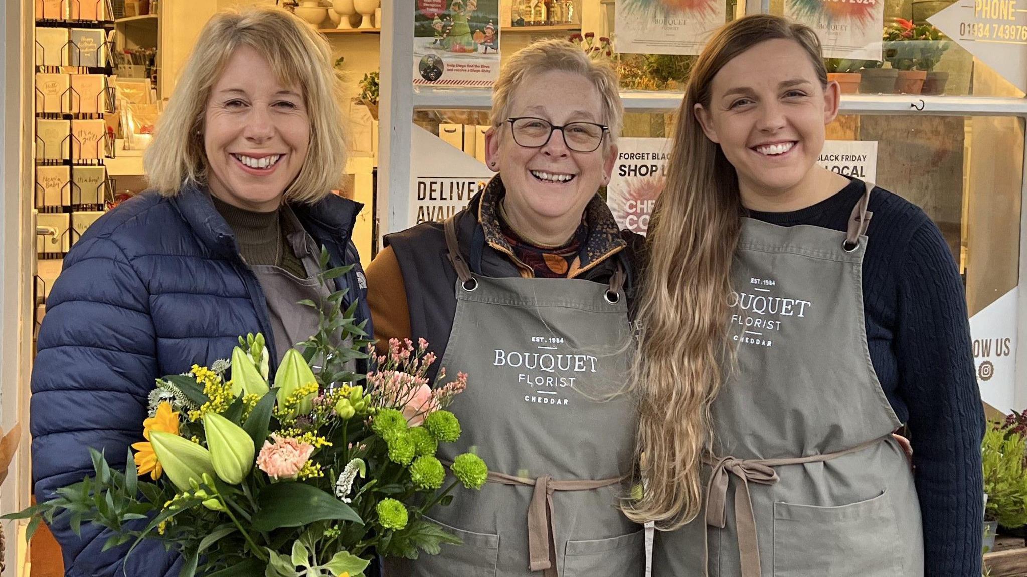 Three women stand outside a florists in Cheddar in Somerset. Two of them are wearing branded grey aprons, and the third is holding a large bunch of flowers with green and light pink blooms.