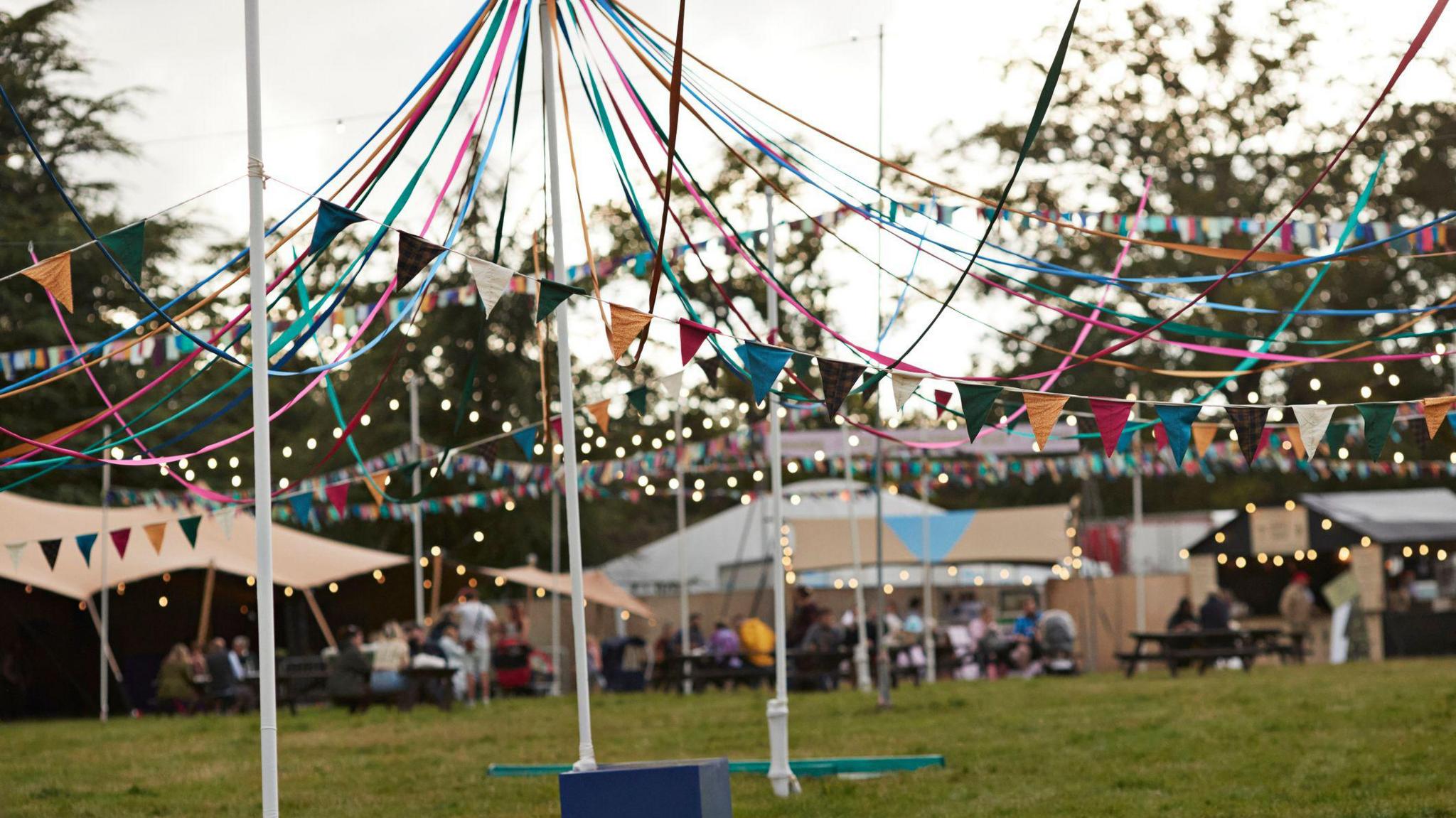 Colourful bunting and fairy lights are strung up between flagpoles  with festival huts and trees in the background.