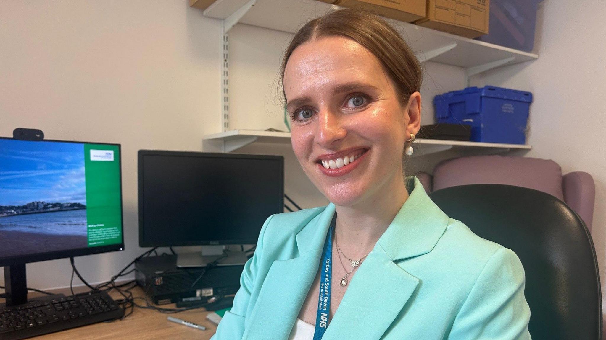 A woman with brown hair and a bright turquoise suit sitting on a chair in an office