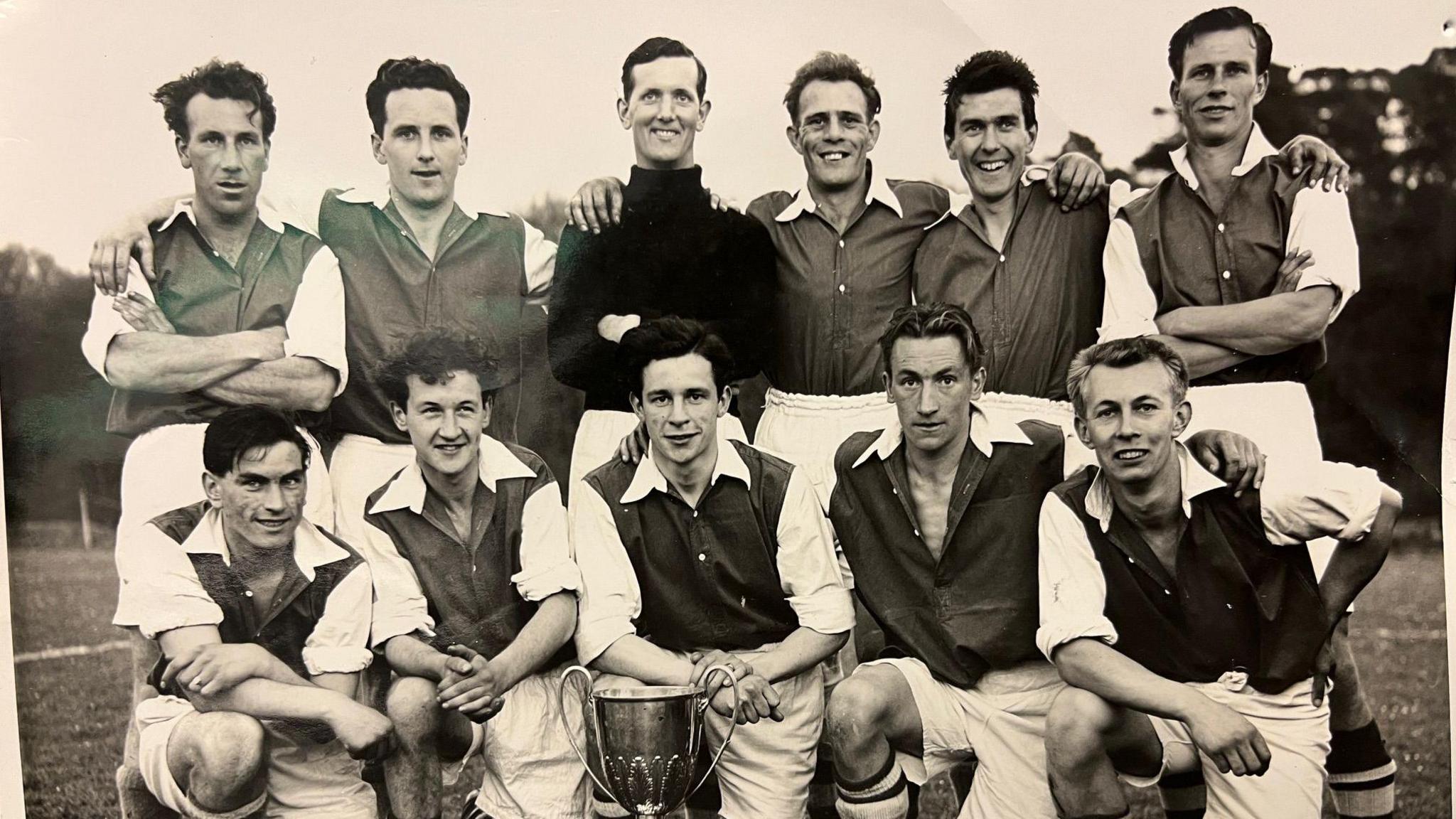 A black and white photo of the Burgess Hill Town Football Club team in 1957. 11 men are posing for the photo with a large trophy at the centre front. 