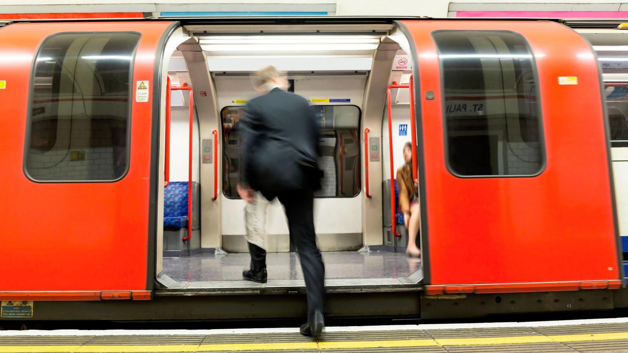 A man in a dark business suit and carrying a newspaper, with his back to the camera, steps on to a London tube carriage.