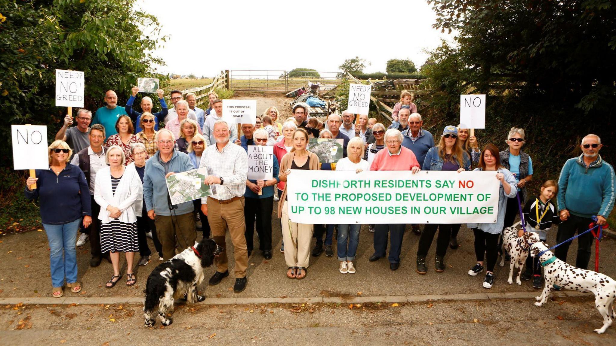 A group of people gathered outdoors, protesting against a proposed housing development. They are holding various signs with messages such as “NO,” “NEED NO GREED,” “ALL ABOUT MONEY,” and a large banner that reads, “DISHFORTH RESIDENTS SAY NO TO THE PROPOSED DEVELOPMENT OF UP TO 98 NEW HOUSES IN OUR VILLAGE.”
The group includes both adults and children, and some individuals are accompanied by dogs. They are standing on a road with greenery and trees in the background.