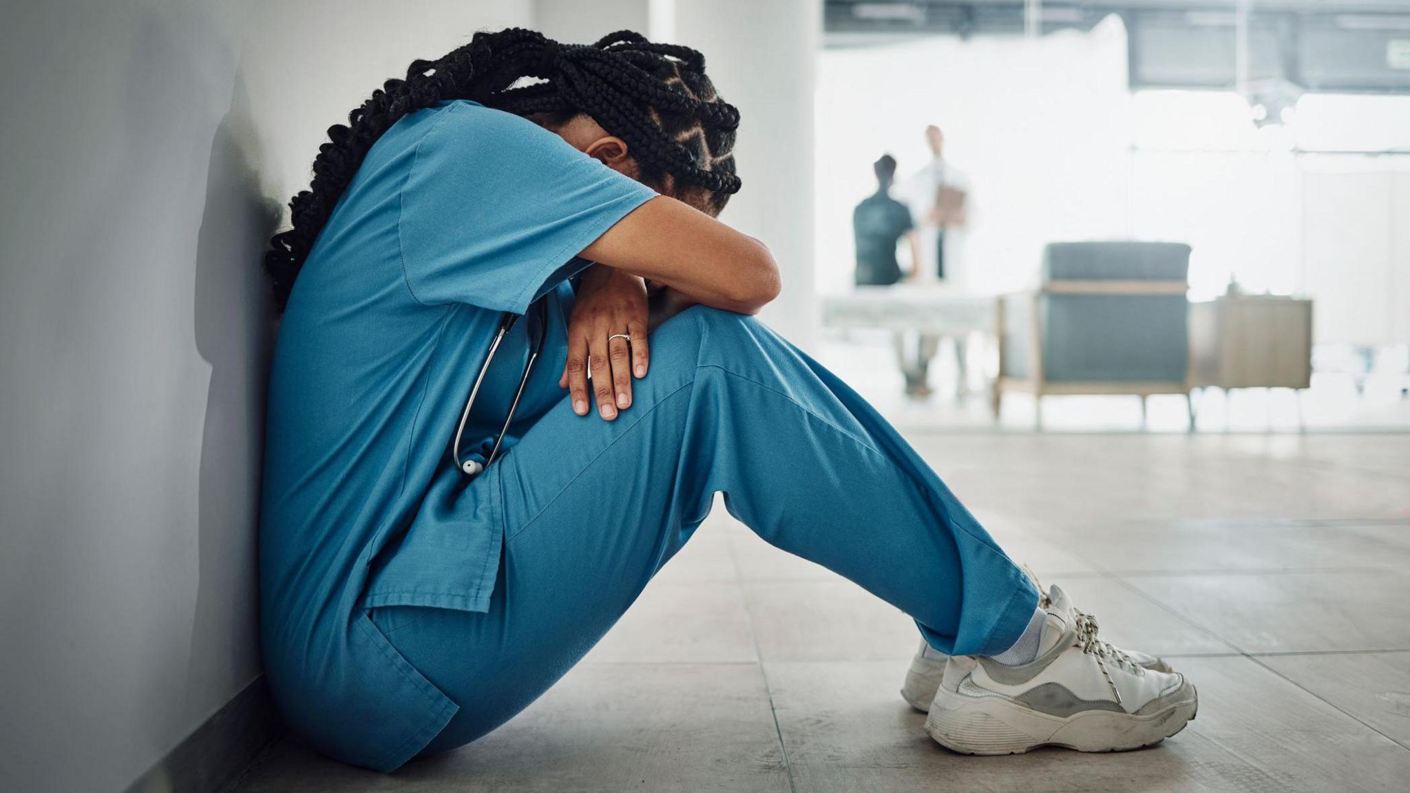 A medic in blue scrubs sits on the floor with head and hands on their knees. They have long dark braided hair and in the background you can see two other people talking in what looks like a hospital environment