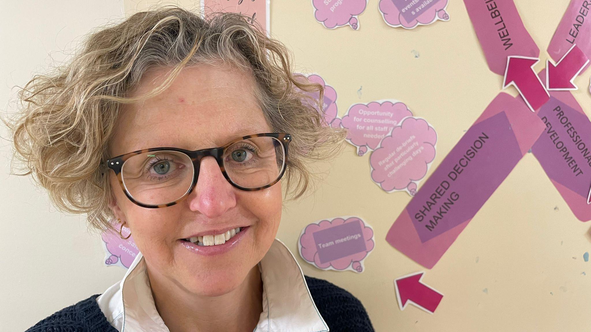 Rachel Westwood with short blonde curly hair. She is wearing tortoise shell patterned glasses and is smiling looking at the camera. Behind her is a display board with pink arrows. 