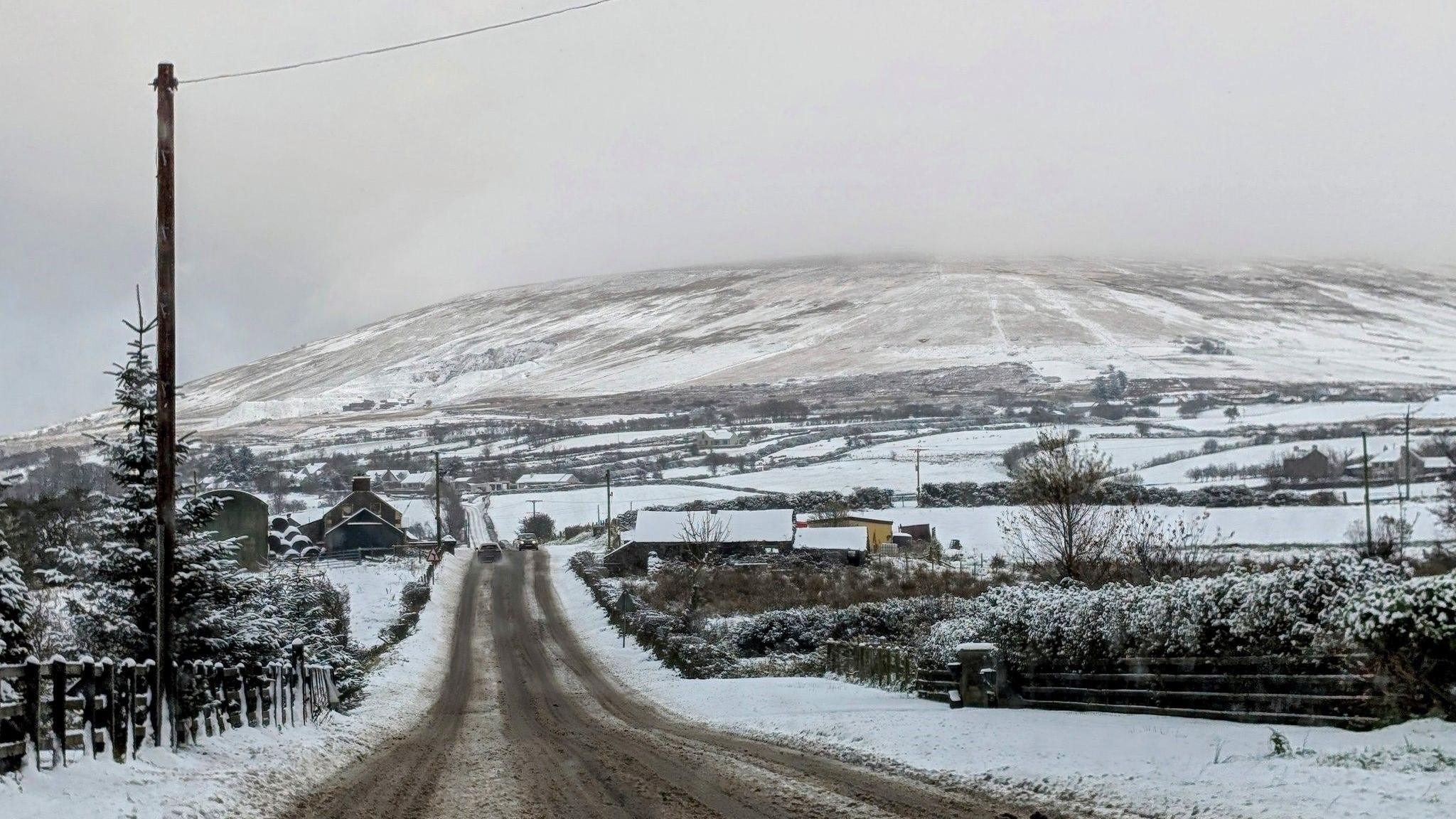 a landscape shot from north antrim in which a road runs up into mountains. the entire scene is snow filled. There are a number of houses along either side of the road, and cars can be seen some distance away