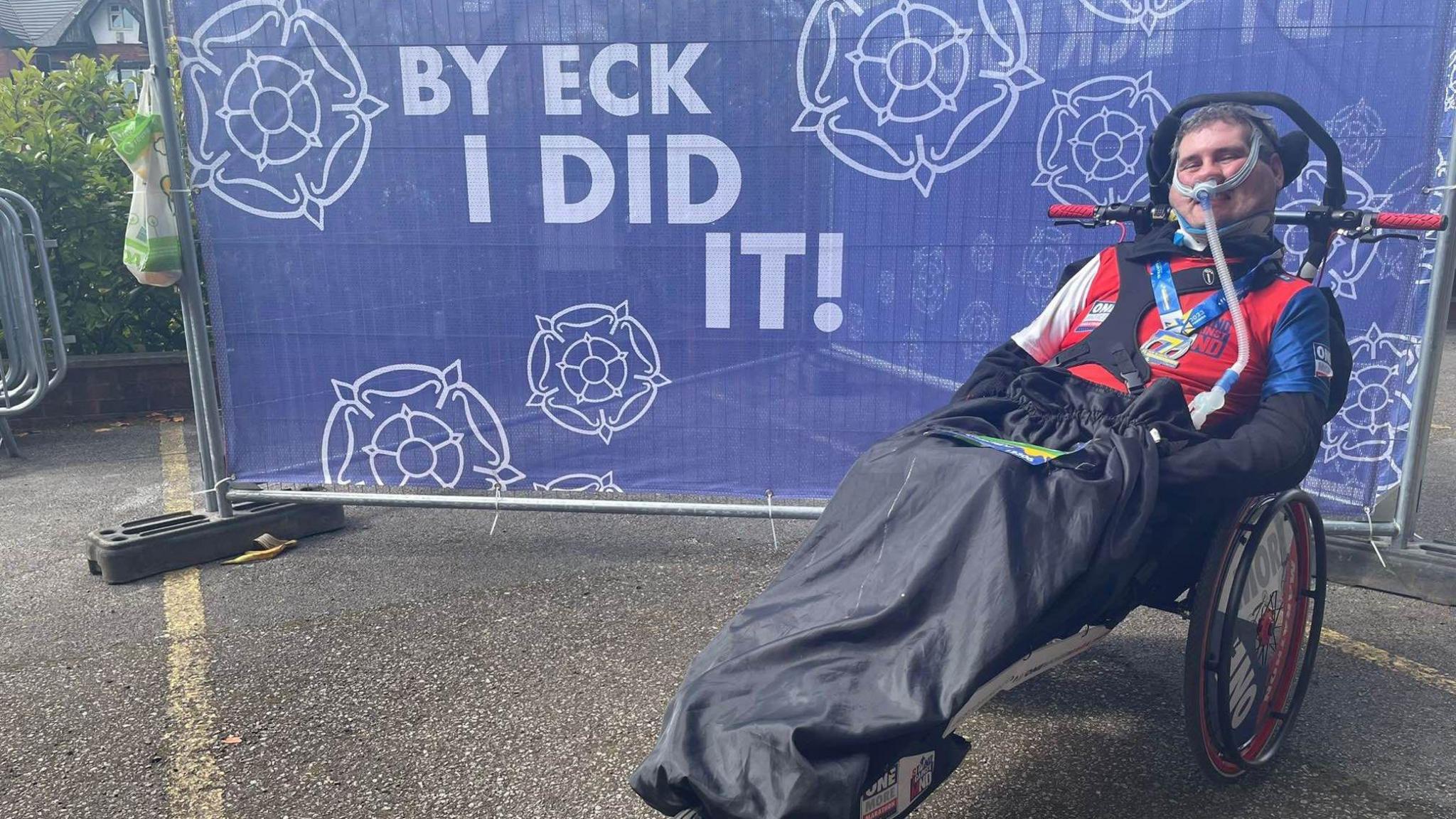 Sam Perkins in a wheelchair next to a sign that reads "By eck I did it"