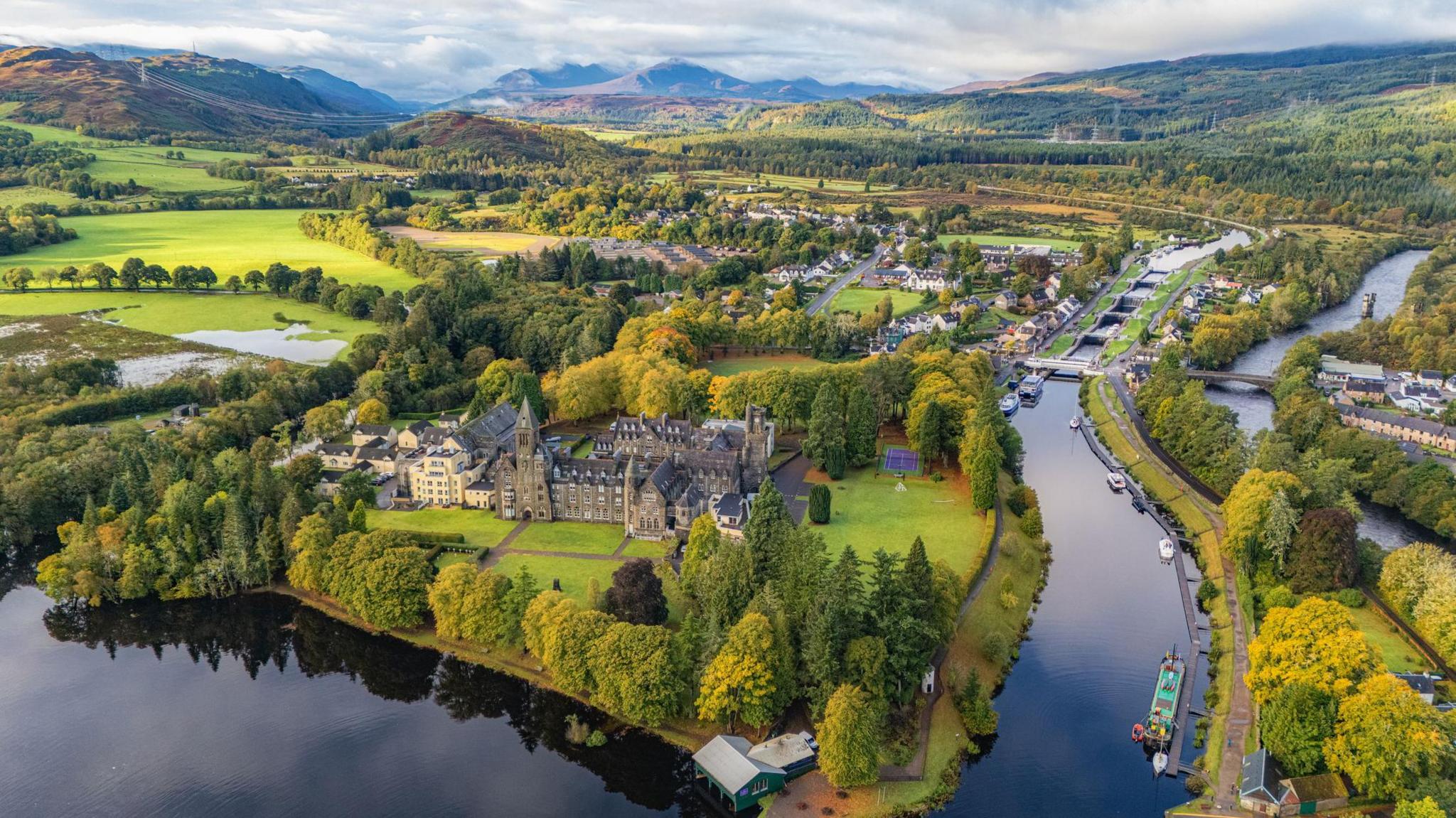 An aerial view of Fort Augustus showing the dark waters of Loch Ness and the Caledonian Canal, and a large abbey among trees on the shores of Loch Ness.