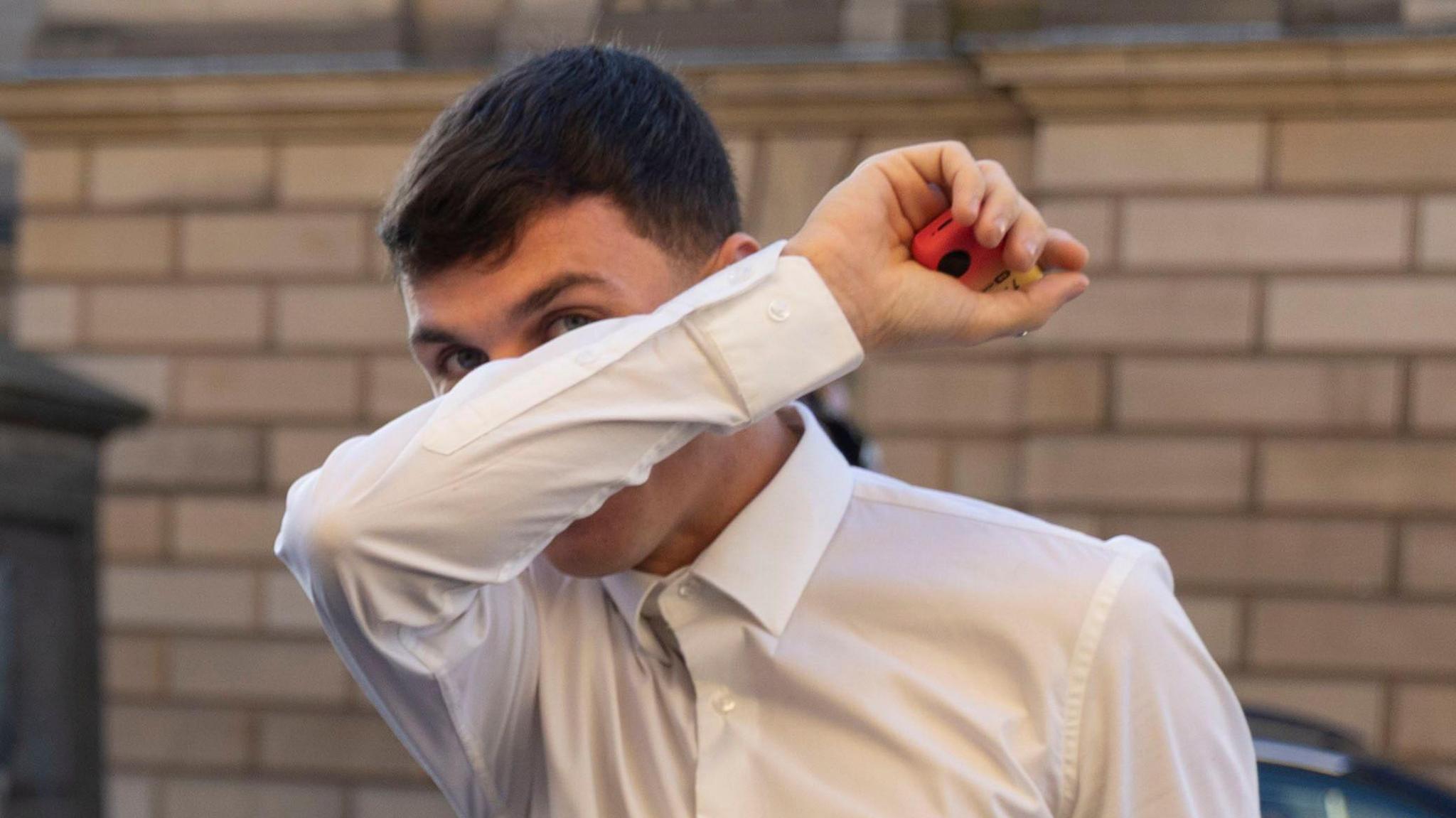 Liam McDermid covering his face with his right arm. He is holding a red and yellow vape in his right hand. He has dark hair and is wearing a white shirt.