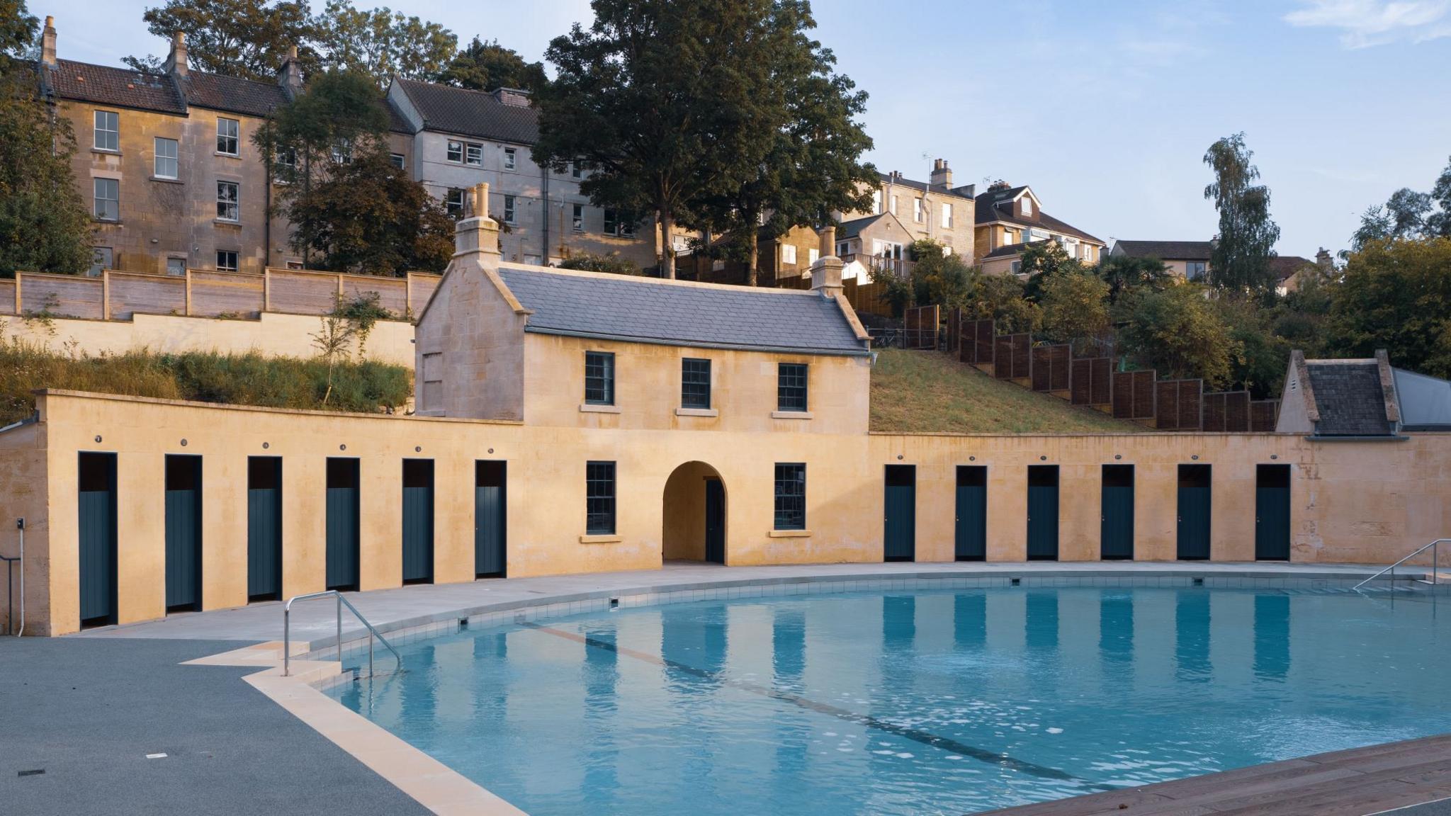 A building curving round a swimming pool at Cleveland Pools in Bath, backed by some houses.