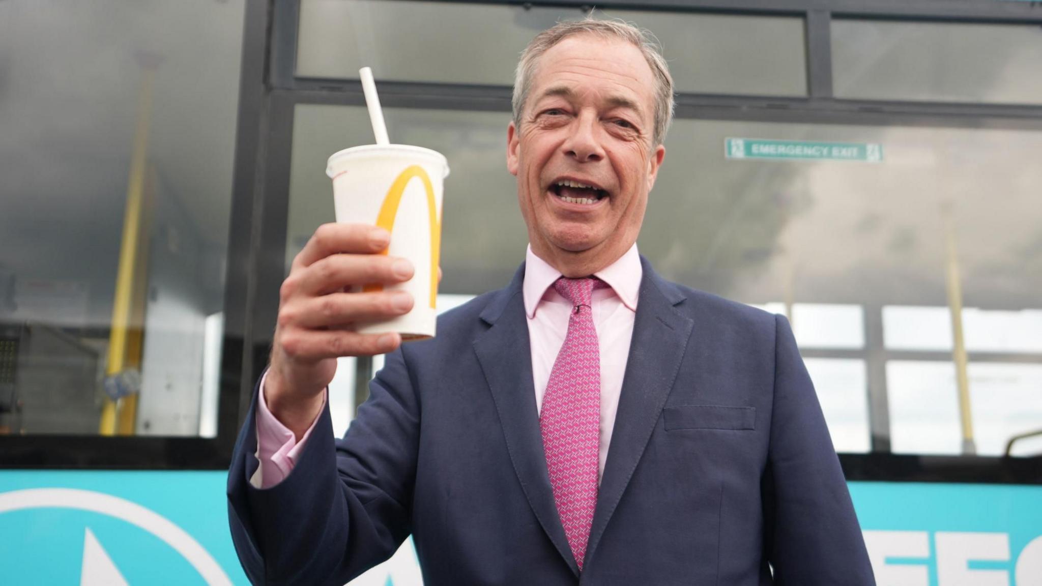 Nigel Farage smiling with his mouth open while holding a McDonald's milkshake. He is wearing a navy suit jacket and pink shirt, with a pink tie.