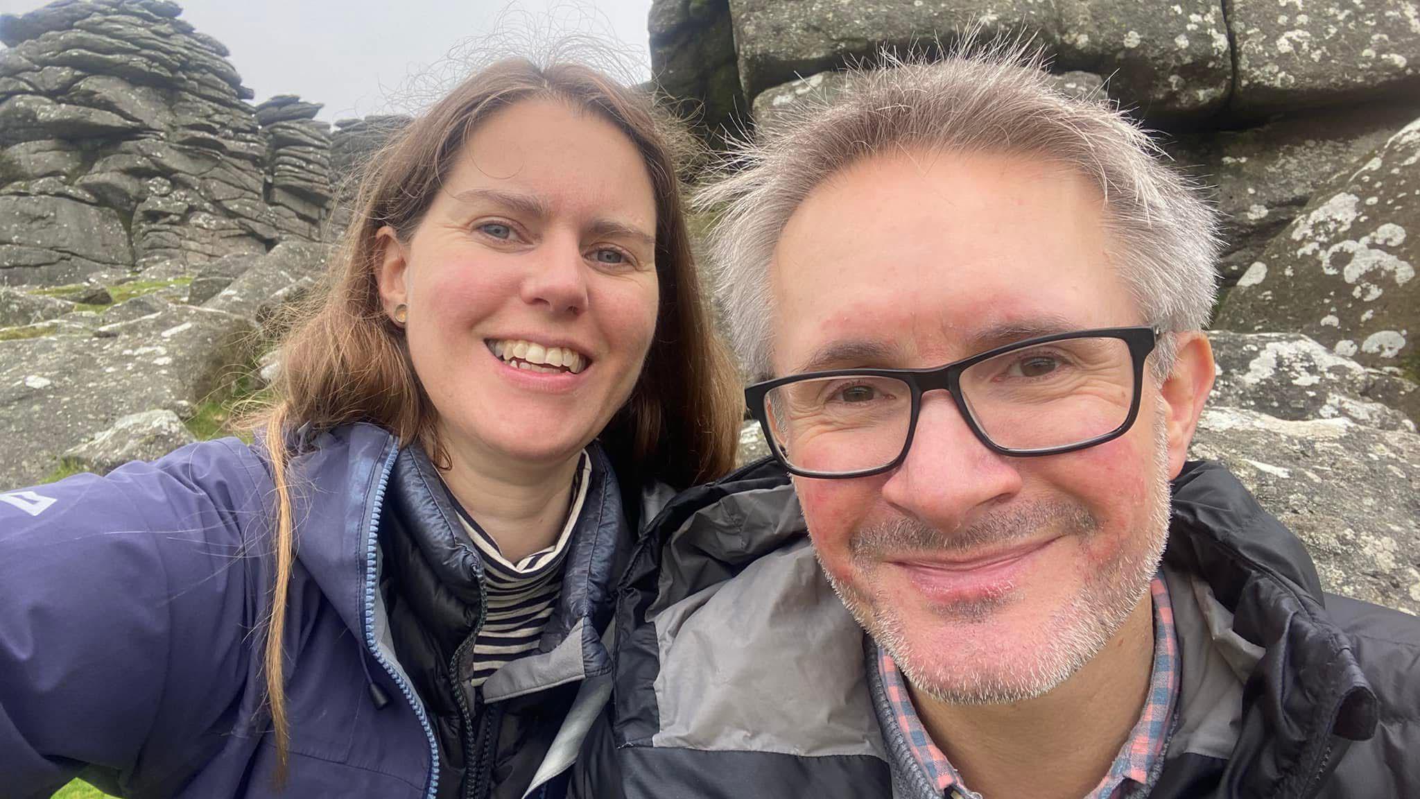 Holly Jackman (left) and Gavin Robbins (right) smile as they take a selfie on Hound Tor in Dartmoor. Holly is stood just behind Gavin and is wearing a purple coat with a black and white stripey top underneath. Gavin has black-rimmed glasses and a black and grey coat.