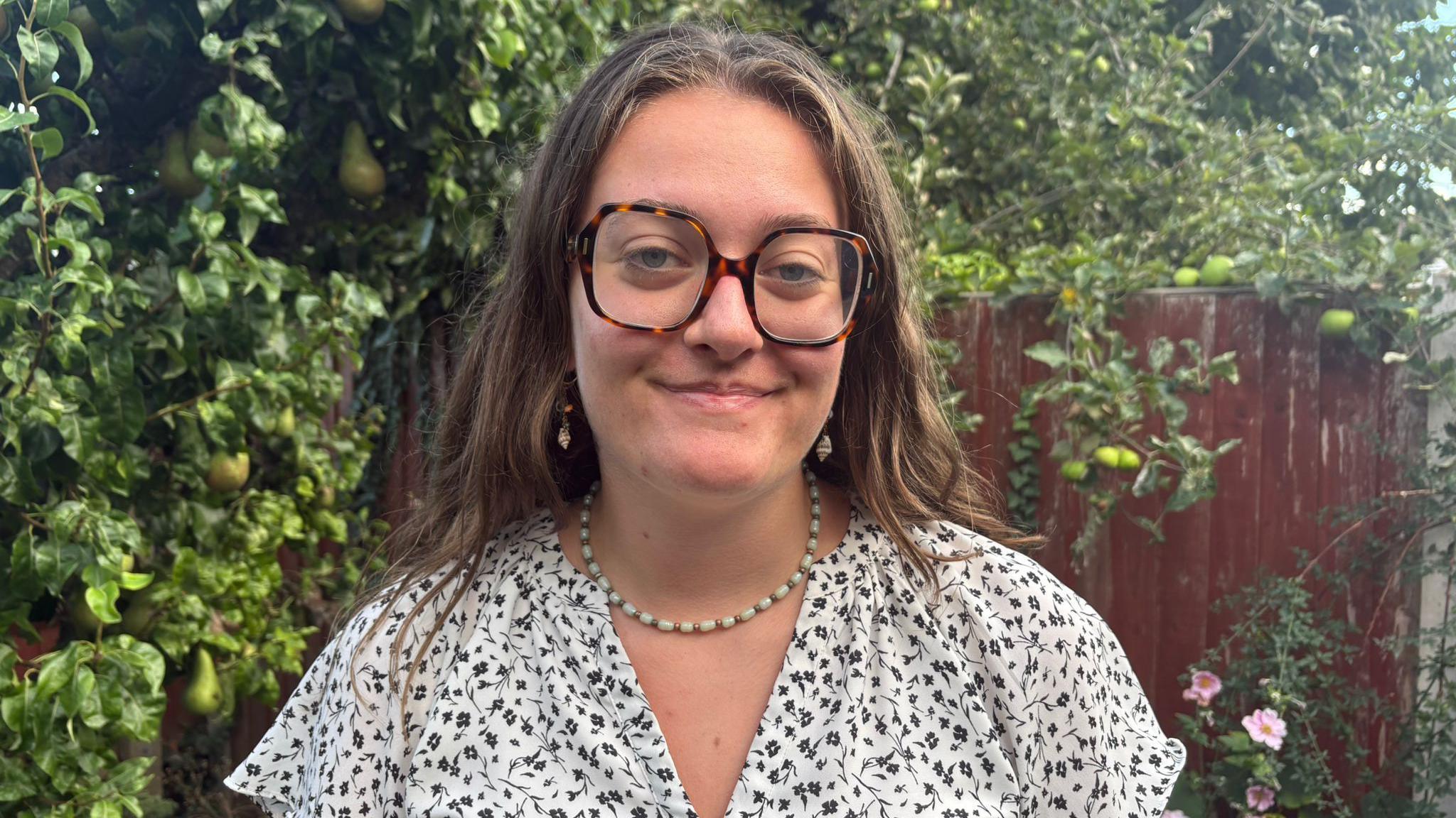A young woman wearing square, tortoiseshell glasses and a white and blue floral shirt smiles while standing in front of a tree laden with pears and a red wooden fence covered in vines.