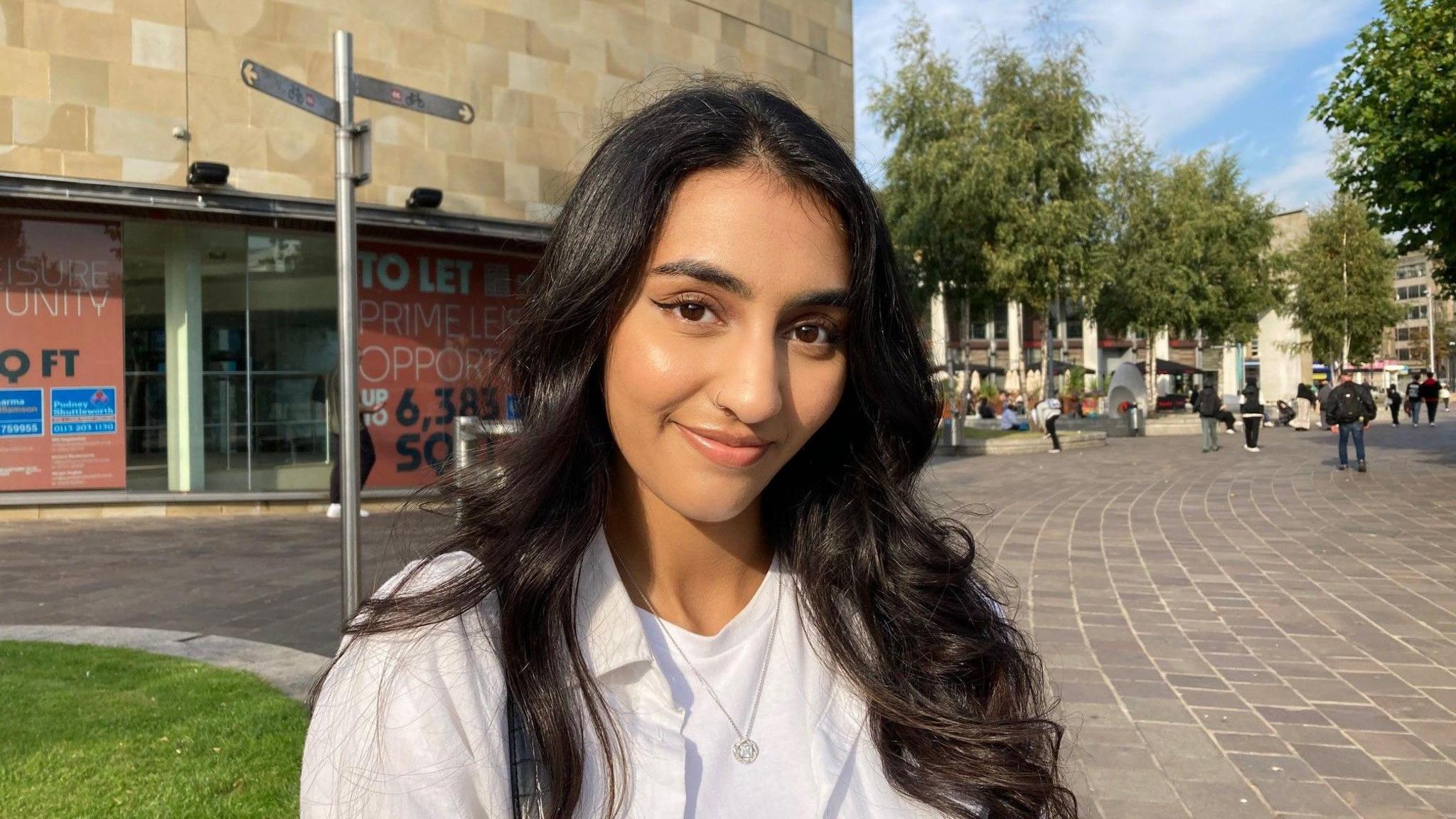 A brown-skinned woman with long, dark hair wearing a white shirt stares at the camera