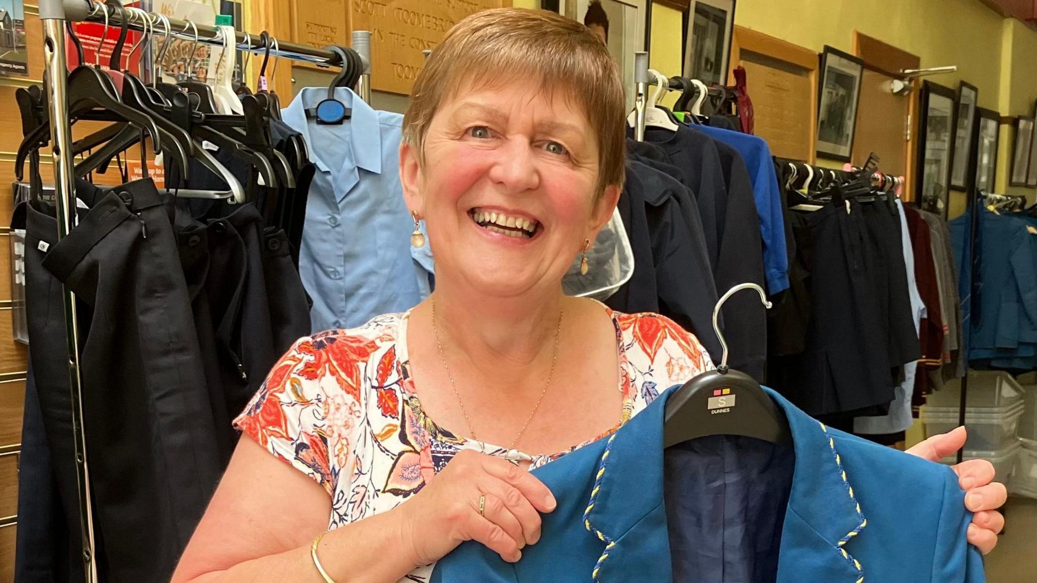 A woman with short light hair wearing a red and white paisley top holds a hanger with a blue school blazer