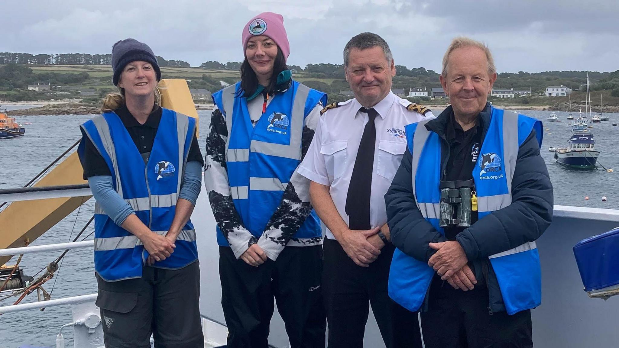  Three members of the ORCA survey team on board Scillonian III. The members are wearing blue ORCA waistcoats. The members are standing in a line, with their arms crossed, and are smiling at the camera. The members consist of one man and two women. Another man, Scillonian III master David Redgrave, is standing next to the members. Mr Redgrave is wearing a white shirt and black tie.
