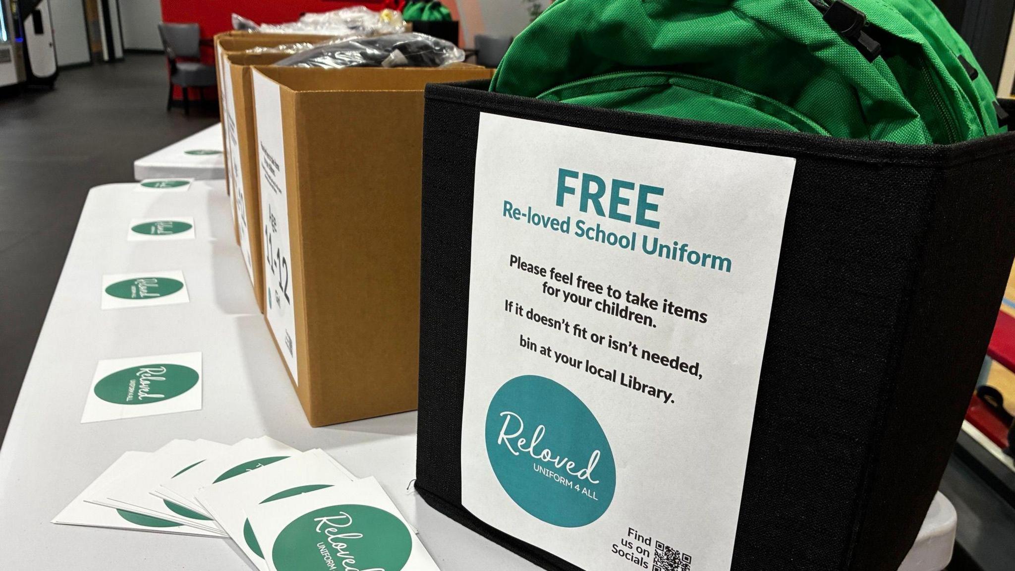 Boxes filled with donated school uniform items are laid out on a table indoors. A sign on the nearest container advertises free "re-loved" school uniform, and a green backpack can be seen among the donated items.