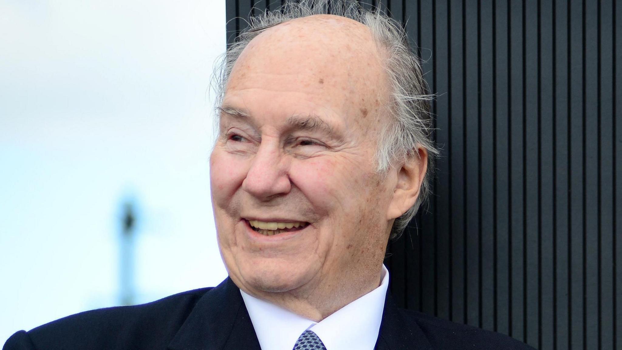 Prince Karim Aga Khan smiling wearing a dark suit, white shirt and blue spotted tie