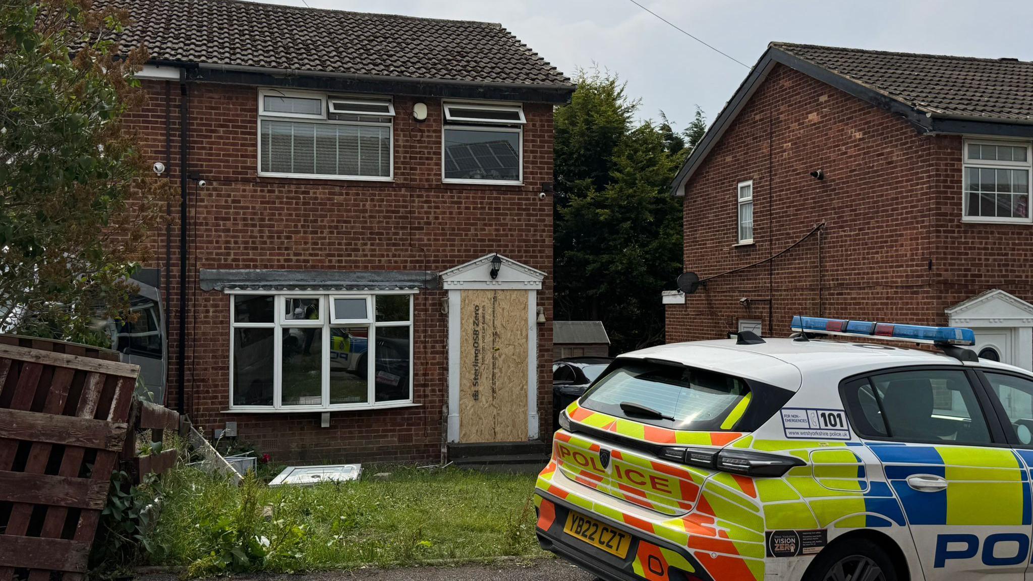 A police car is parked in front of a semi-detached house, which has a boarded up door. 