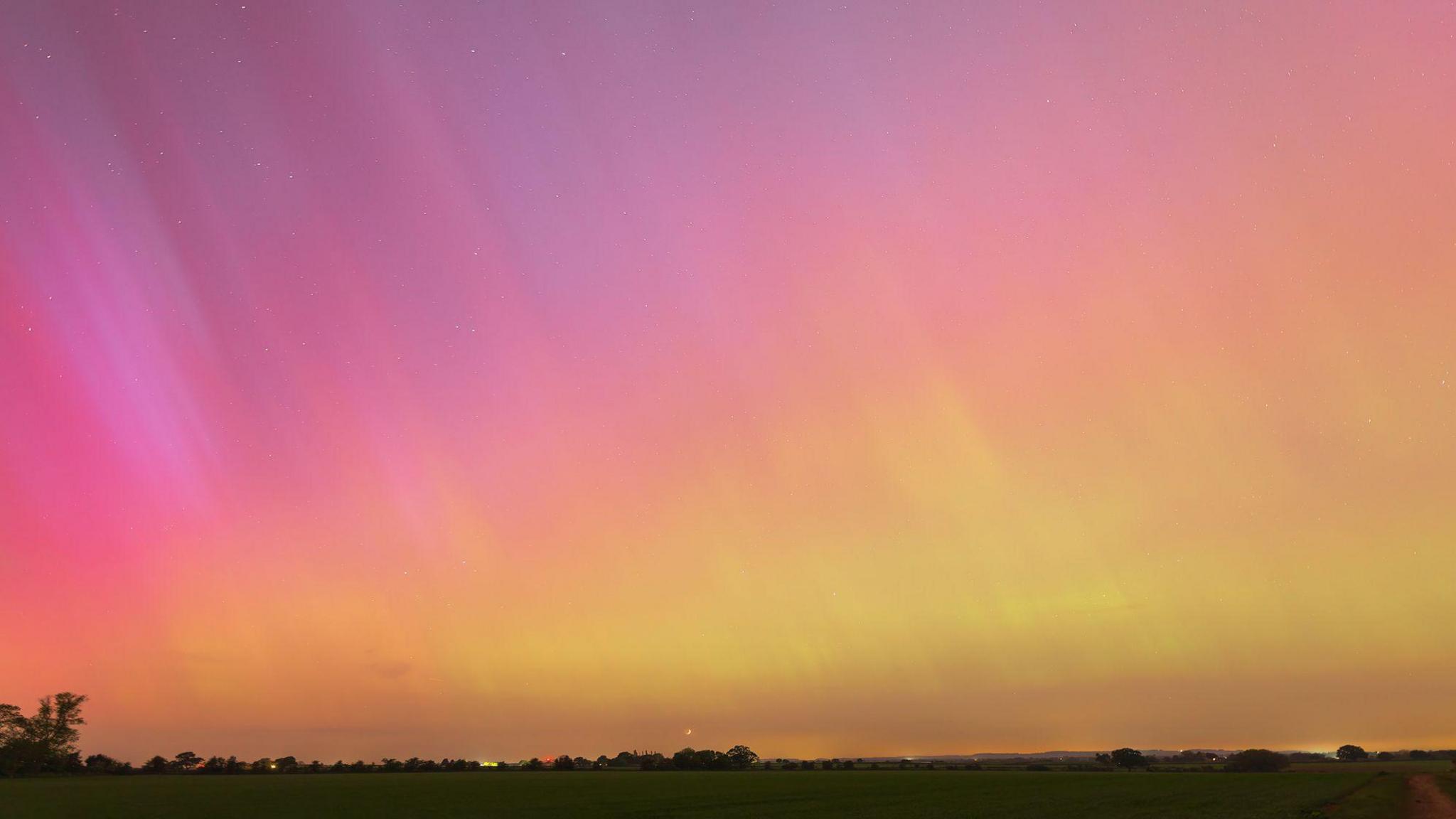 The northern lights pictured from a field. The sky is purple and pink at the top of the image and yellow and orange towards the bottom. The outline of a field, with houses in the distance fills the bottom of the image. Stars can also be seen in the sky. 