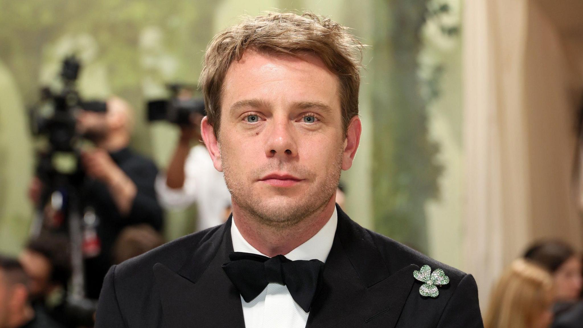 Jonathan Anderson pictured here at the 2024 Met Gala.
He is wearing a black dinner suit with black bow tie and jewelled shamrock on his left lapel/
He is looking directly at the camera and has fair short hair and has a bit if stubble.
Behind him you can see cameras.