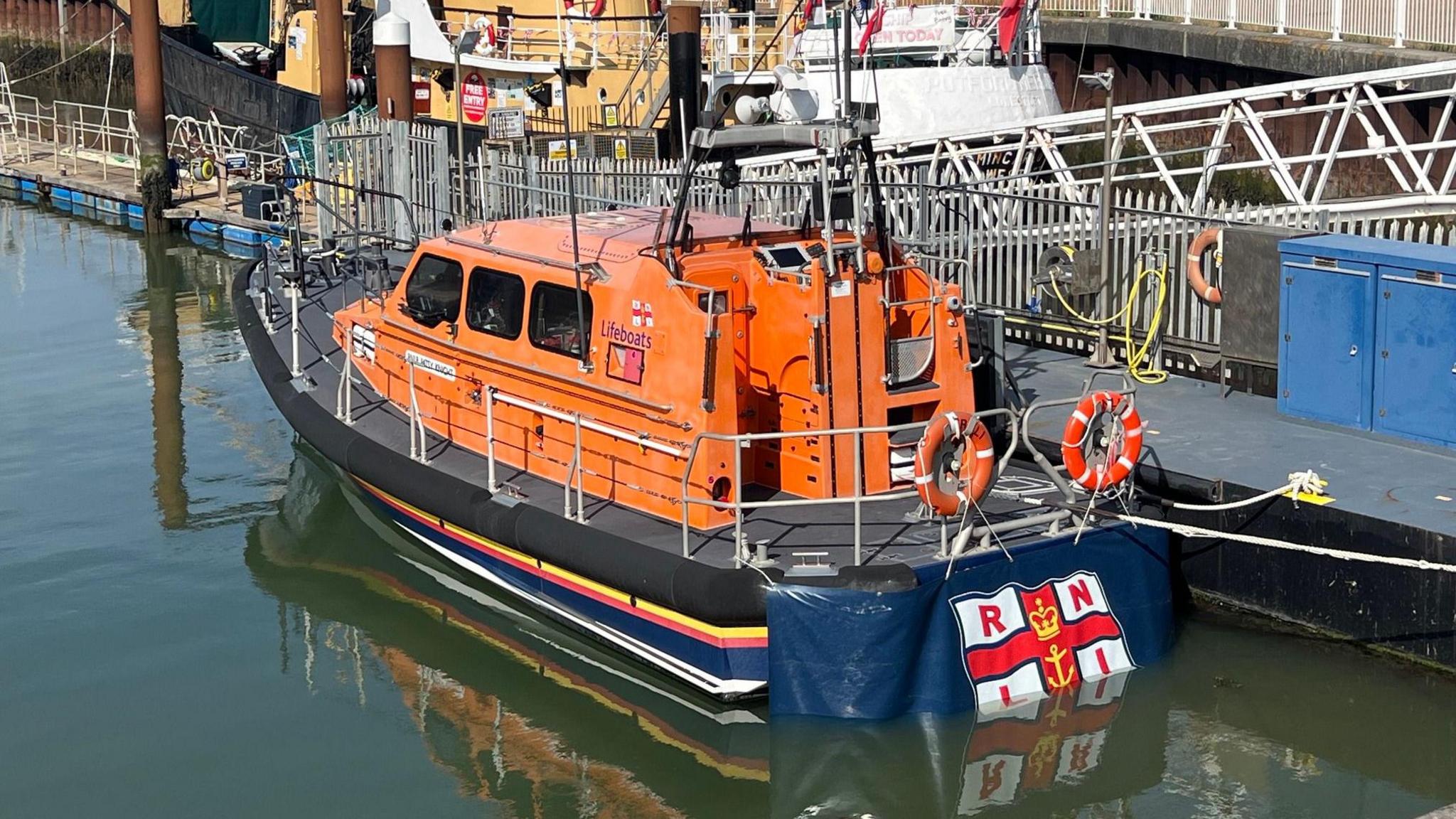 An orange lifeboat is moored next to a dock. It has an RNLI flag on the rear of the vessel. 