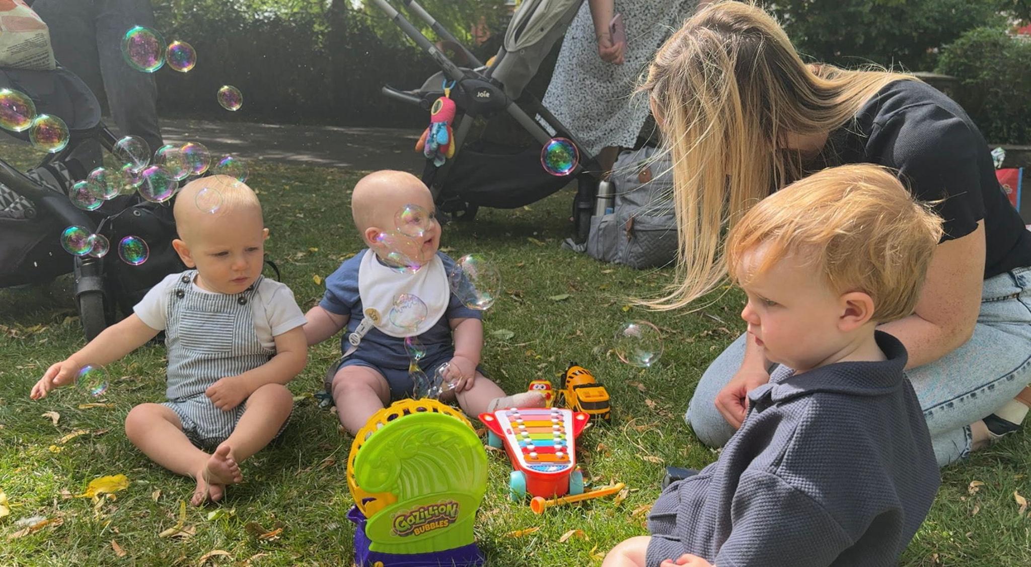 Two babies sitting on the grass. One is wearing blue-striped dungarees, the other a blue outfit with white bib. They are looking at a blond-haired woman who is kneeling down. In the foreground, a toddler wears a blue shirt and looks at a green bubble machine. There is a colourful xylophone on the grass.