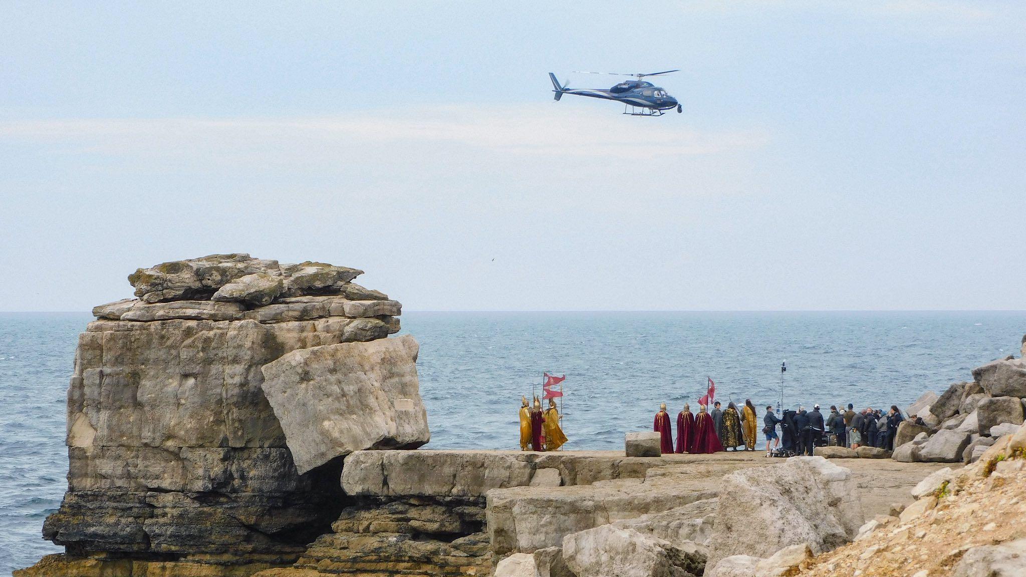A helicopter flying over Pulpit Rock while actors and crew shoot a scene underneath by the sea. The weather is overcast.