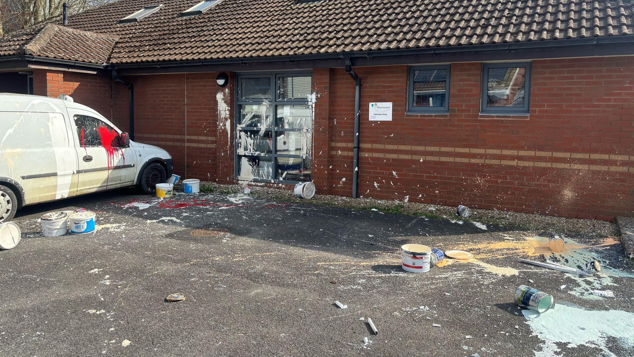 Almost a dozen cans of paint can be seen strewn across the floor outside a therapy centre. Splotches of blue, orange and white paint appear to have been thrown across the car park and building's windows. A white van has also been coated in white and red paint.