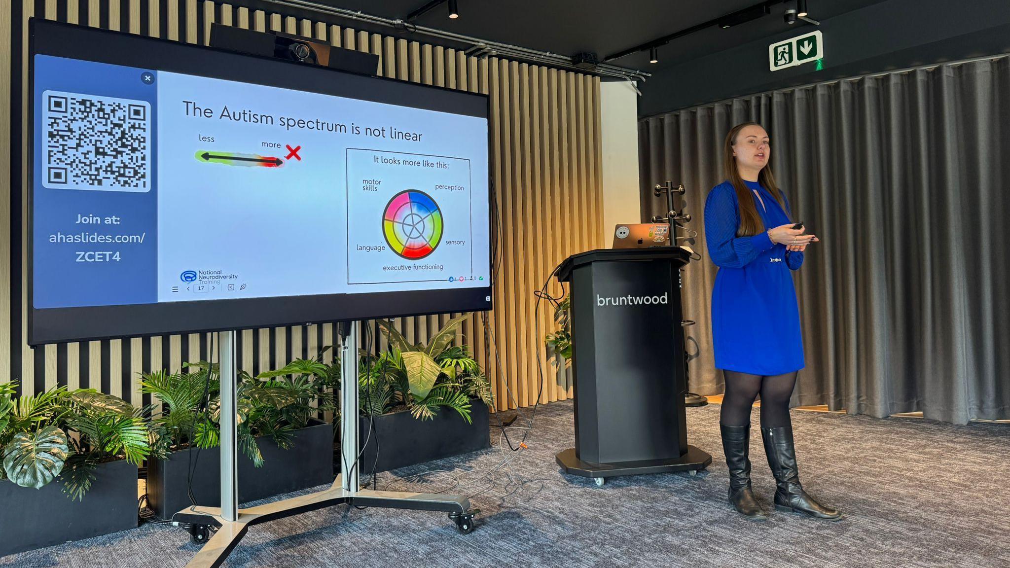 A woman wearing a blue dress stands and delivers a presentation.