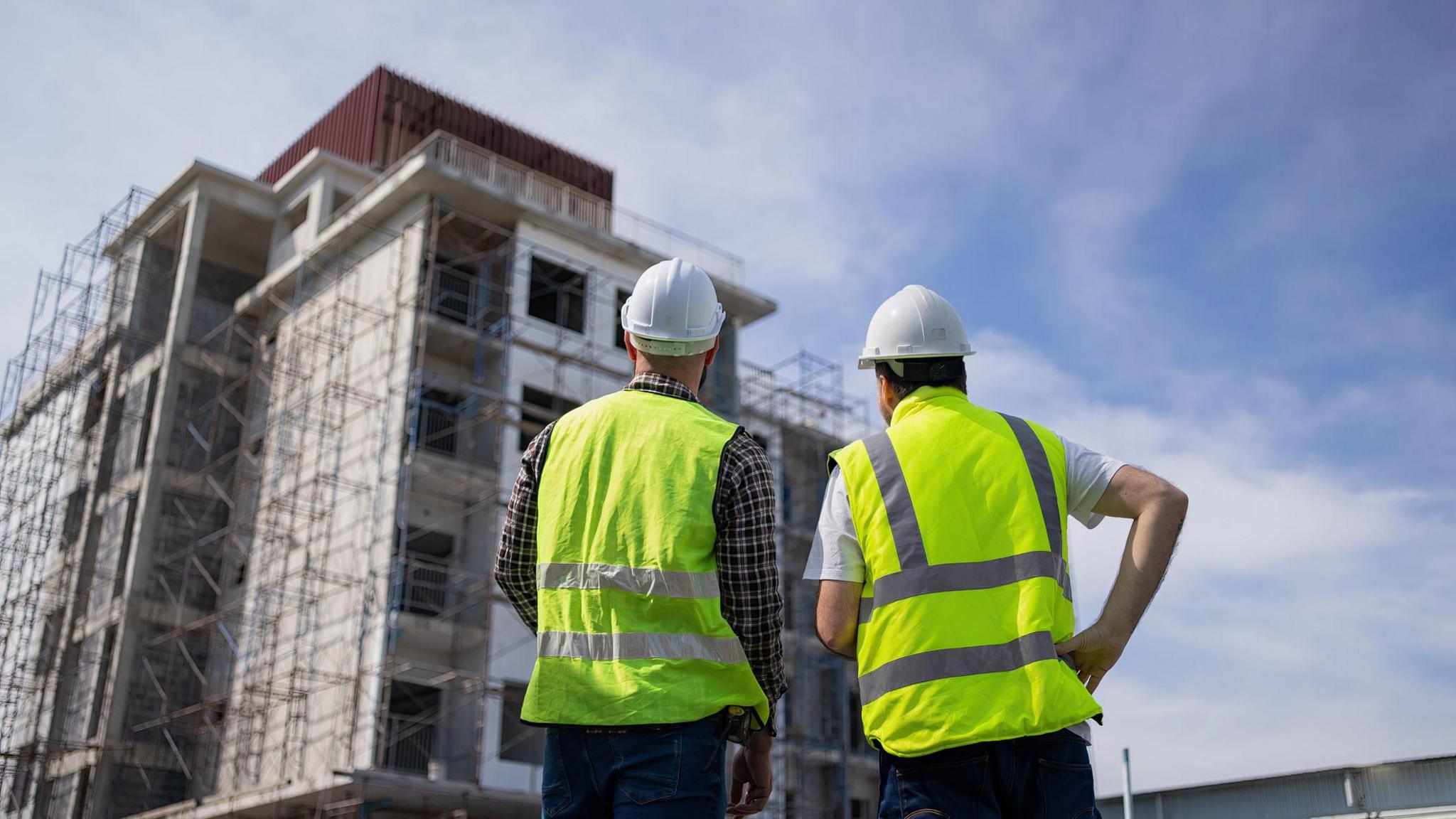 Two men in high visibility jackets look up at a building under construction