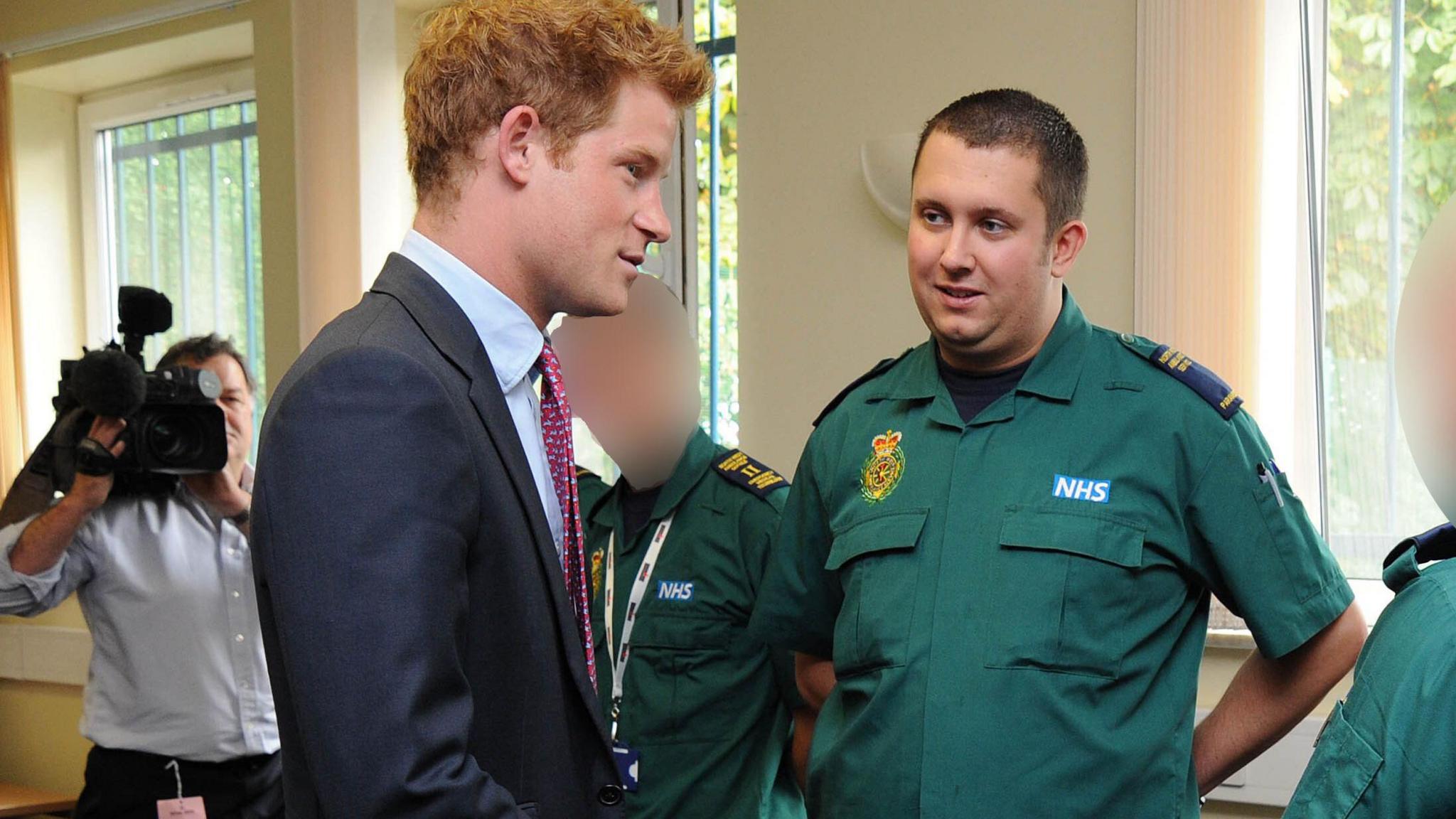 Jonathan Kirk, who has short dark hair and is wearing a green paramedic uniform, stands in front of Prince Harry, who has ginger hair and is wearing a navy blue suit and burgundy tie. 