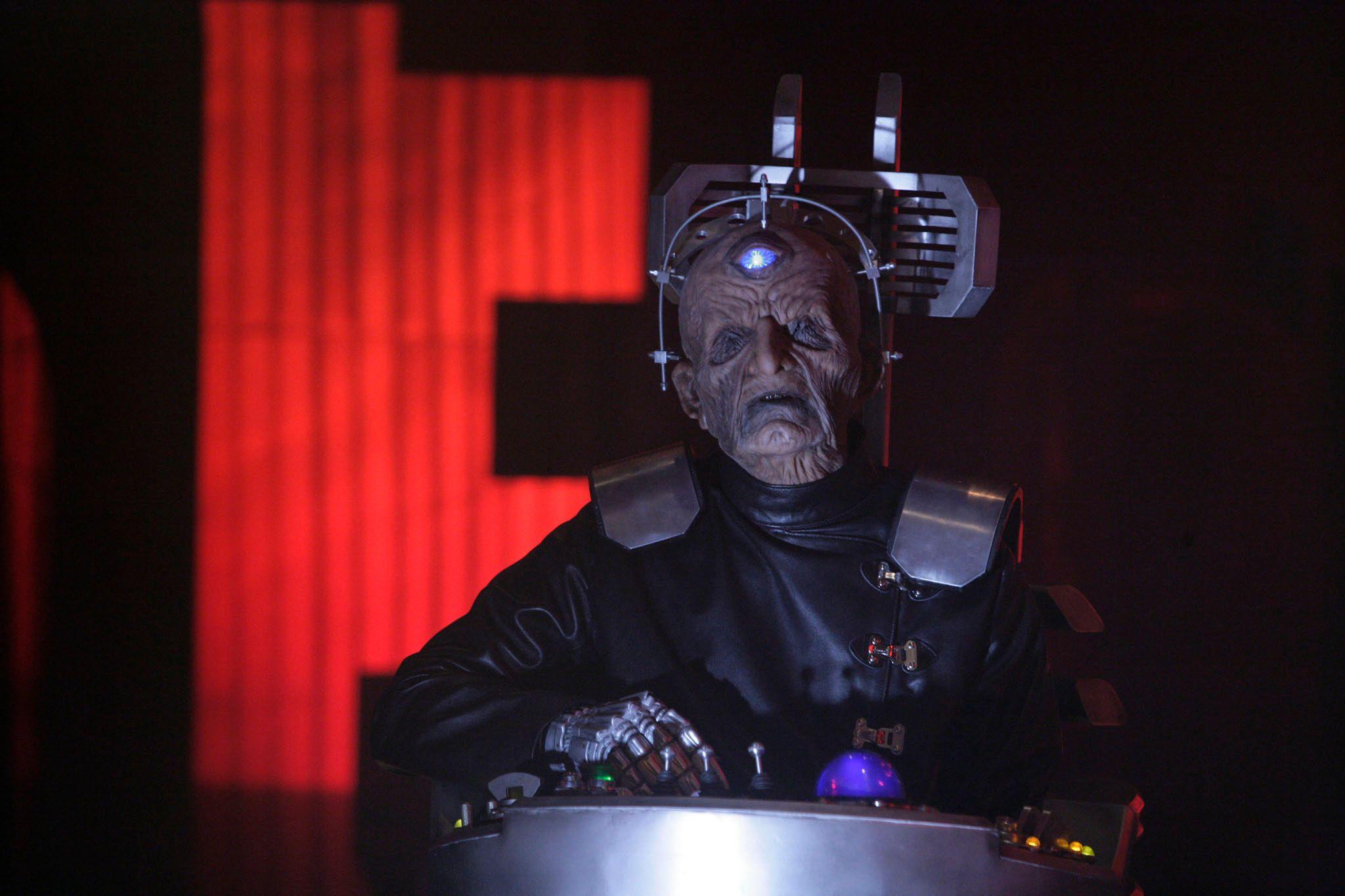 A shadowy figure of Davros in his life-support chair with his robotic hand on switches