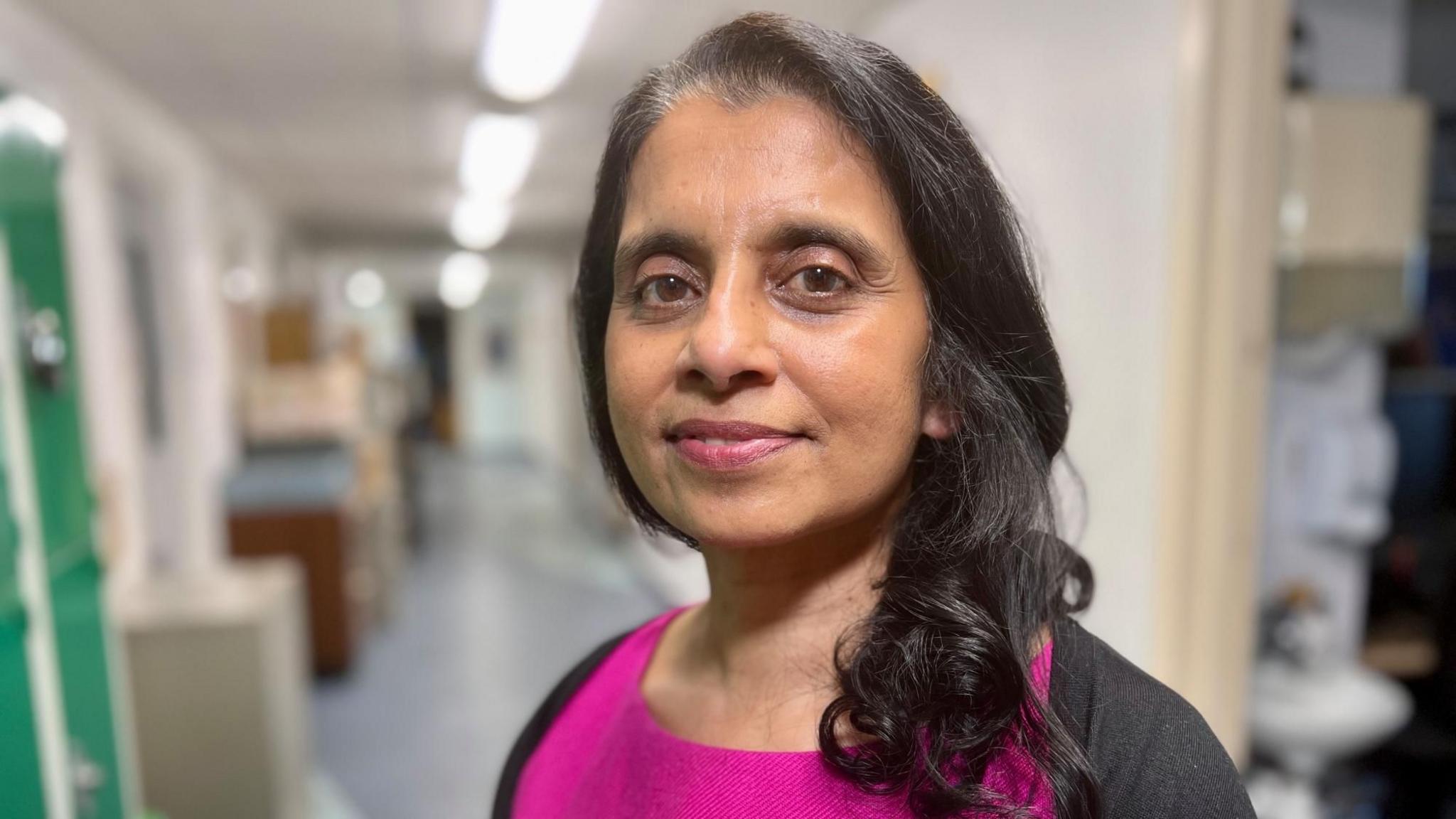 Professor Jean Abraham looks at the camera and is standing in a hospital corridor. She is wearing a pink dress, black cardigan and has black hair.