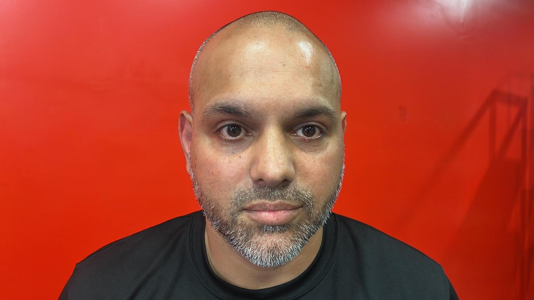 Bilal Javed is looking towards the camera with a light smile. He has a short grey beard and brown eyes, and is wearing a black T-shirt. He is standing in front of a bright red background.