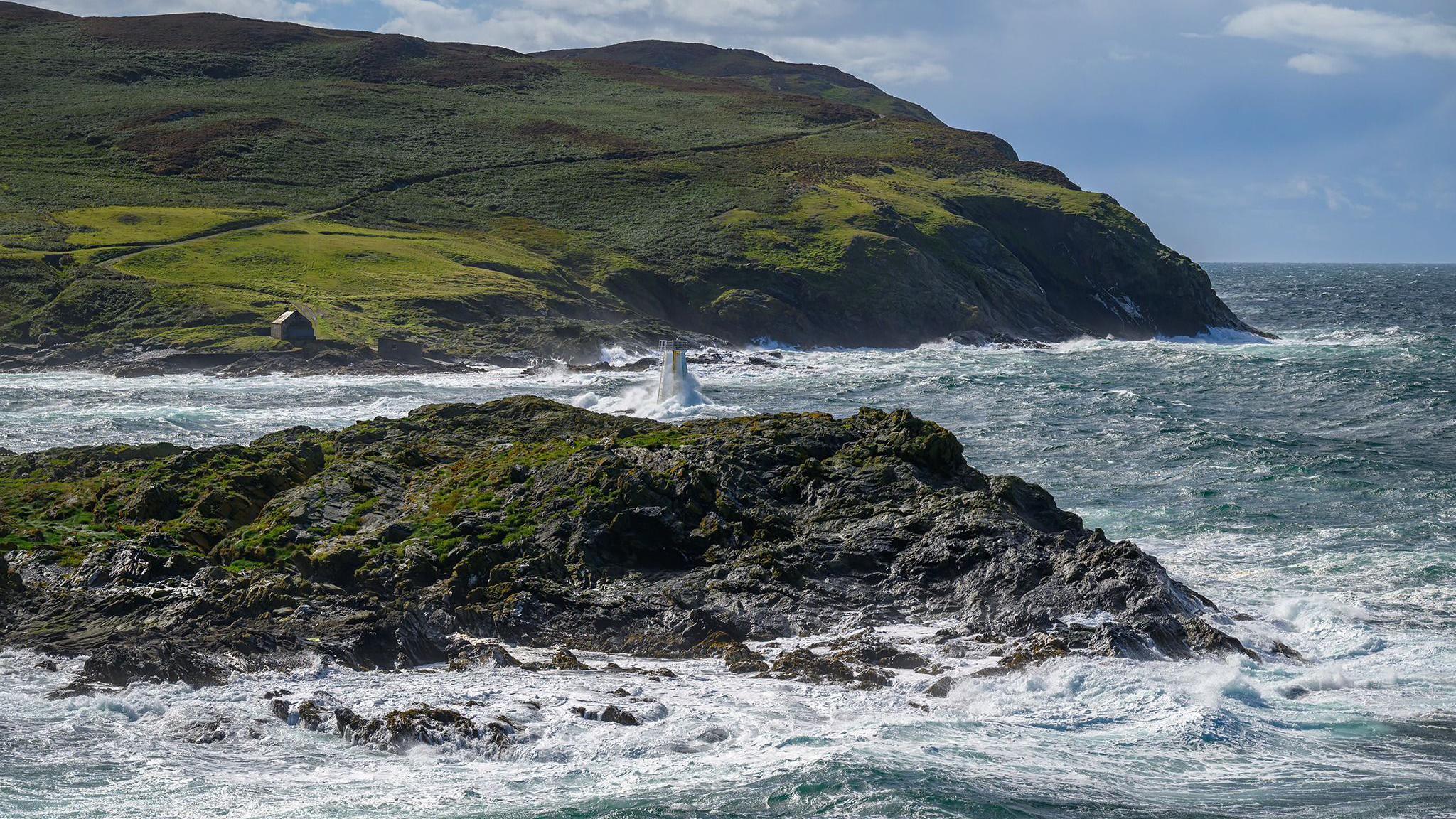 Chicken Rock, a rocky islet off the coast of the Isle of Man, on a windy day with the sea crashing against the rock creating a white spray. In the background is a green hillside on the Calf of Man. 