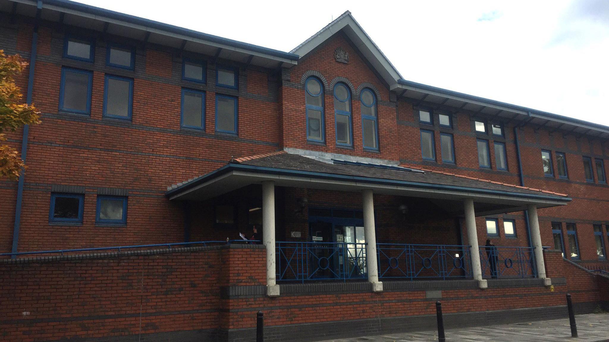 Outside of Stoke-on-Trent Crown Court. It is a modern brown building with blue window frames and sloped path going up to the entrance. 