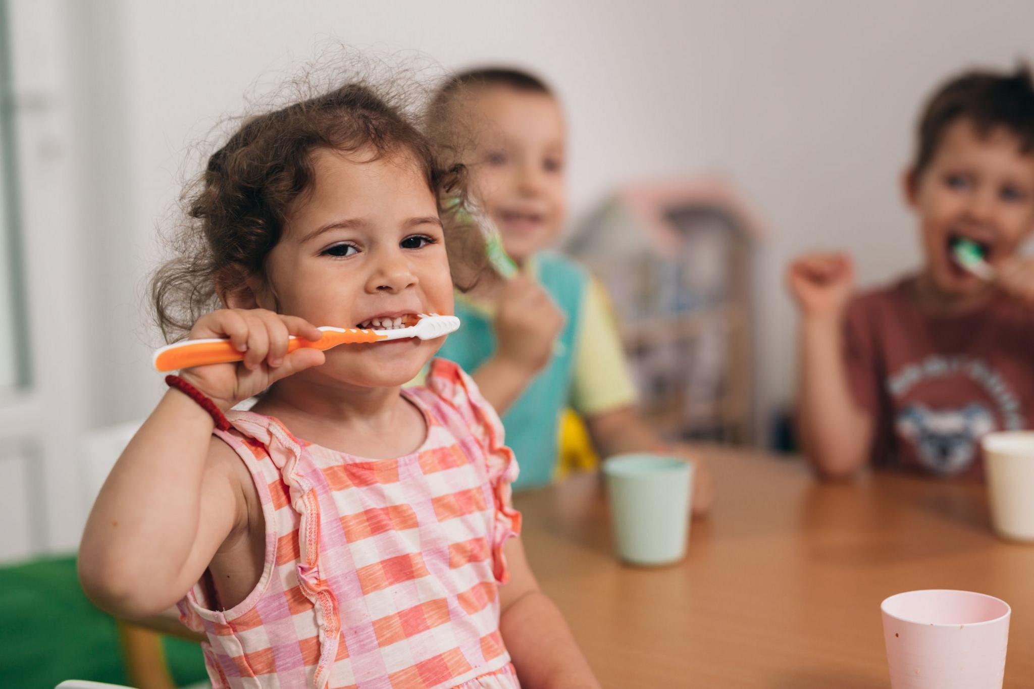 Young children sat around a table brushing teeth. Image focuses on young girl with curly grown hair and striped peach tshirt, smiling as she brushes her teeth. There are pastel coloured plastic beakers on a wooden table and two young boys in the background who are also brushing their teeth