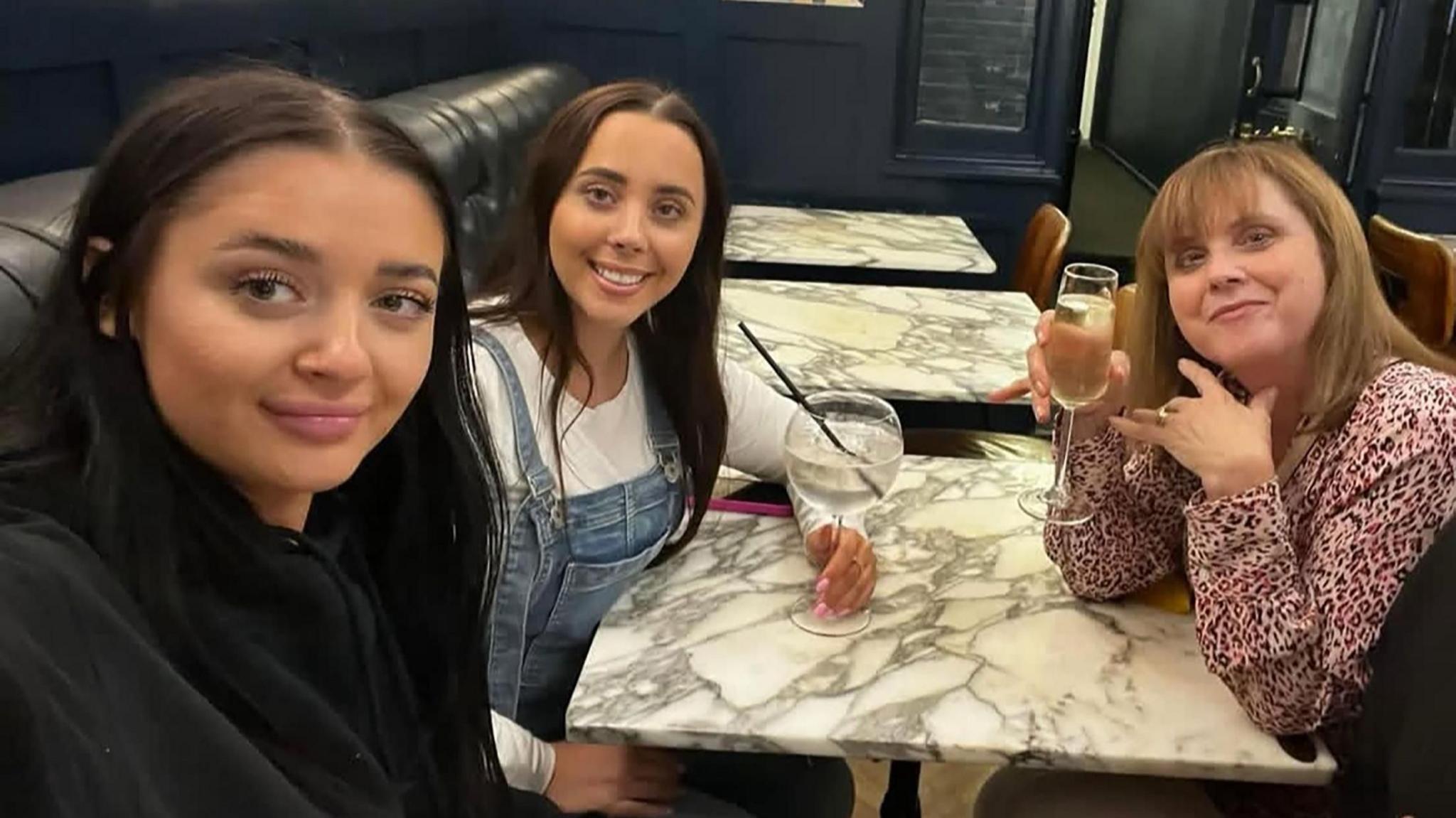 An image of Carol, Louise and Hannah Hunt sitting in a restaurant and smiling. From left to right, Louise has long black hair, is wearing black, Hannah has long dark hair and is wearing a white top under blue dungarees. Carol has light brown hair and is wearing a pink top with a small dark print.