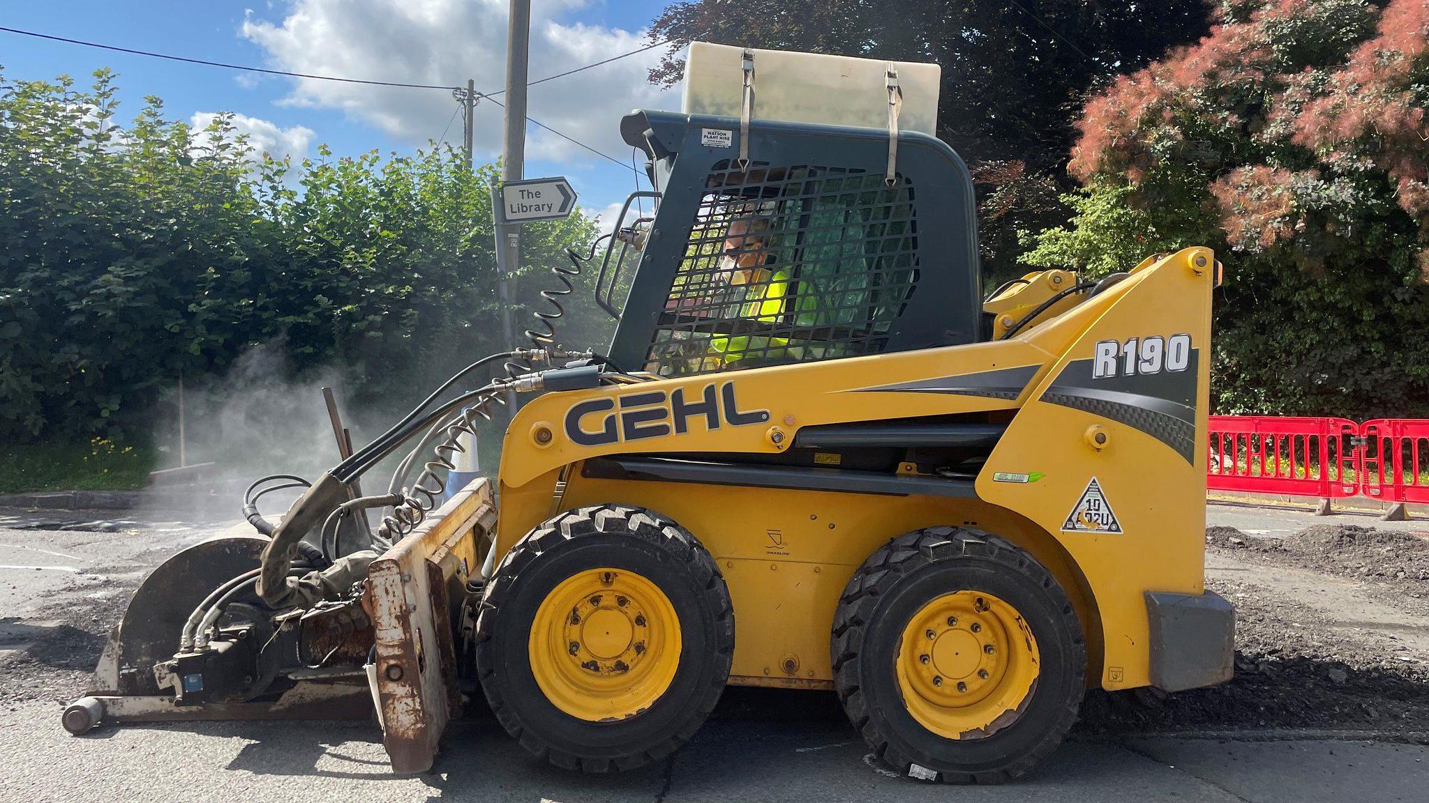 Small yellow vehicle with tractor tyres, a front attachment for folling potholes, and a caged cab for the driver