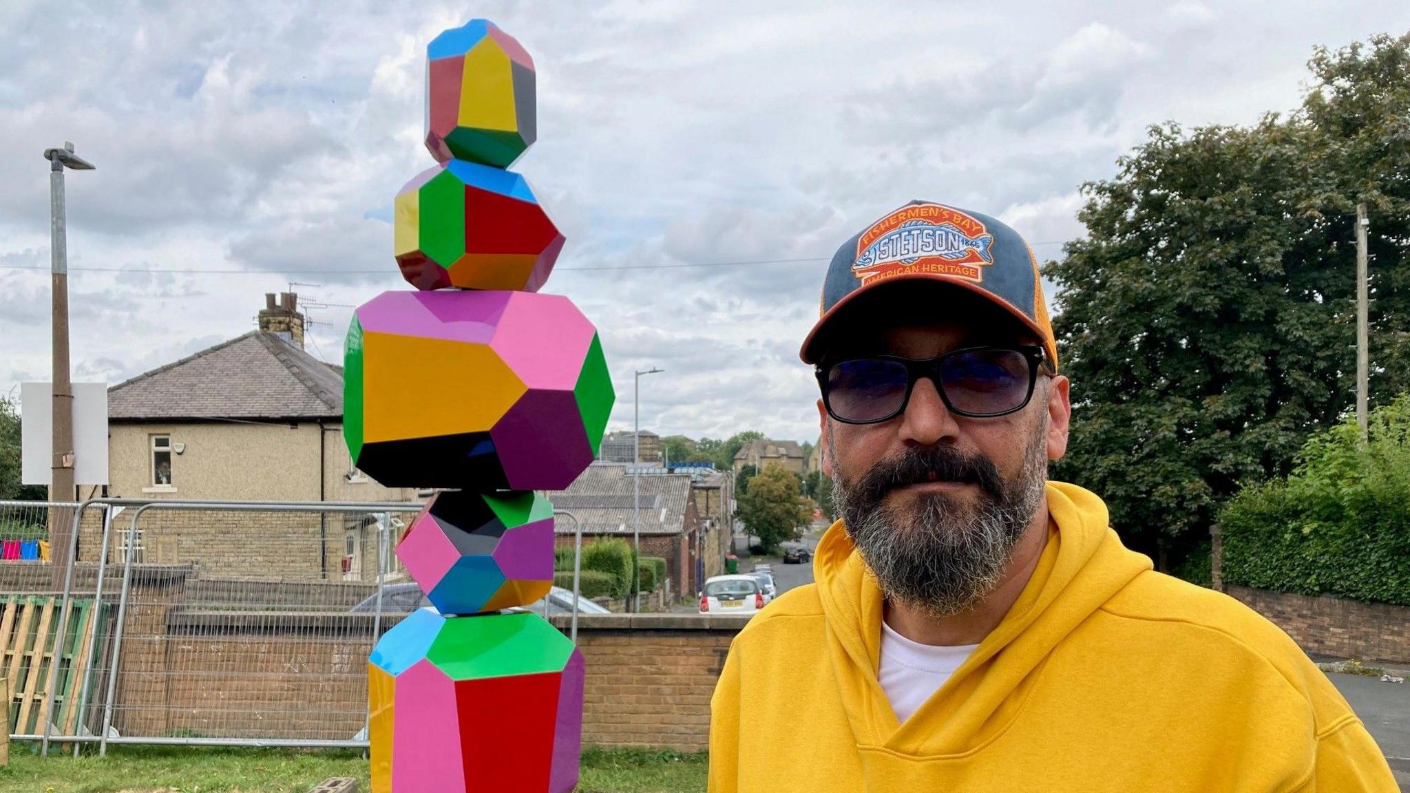 A man in sunglasses, a baseball cap and yellow hooded top looking directly at the camera with a multi-coloured sculpture in the background.