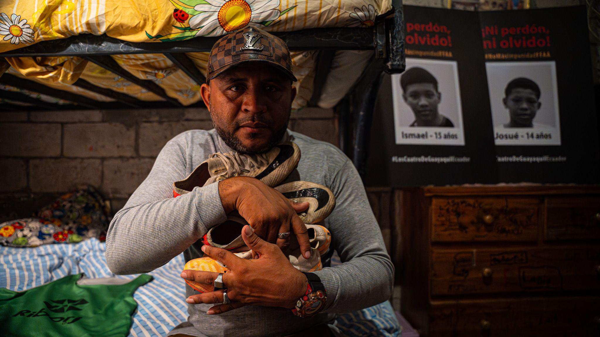 A man is seen holding football boots, with posters of his two teenage sons behind him