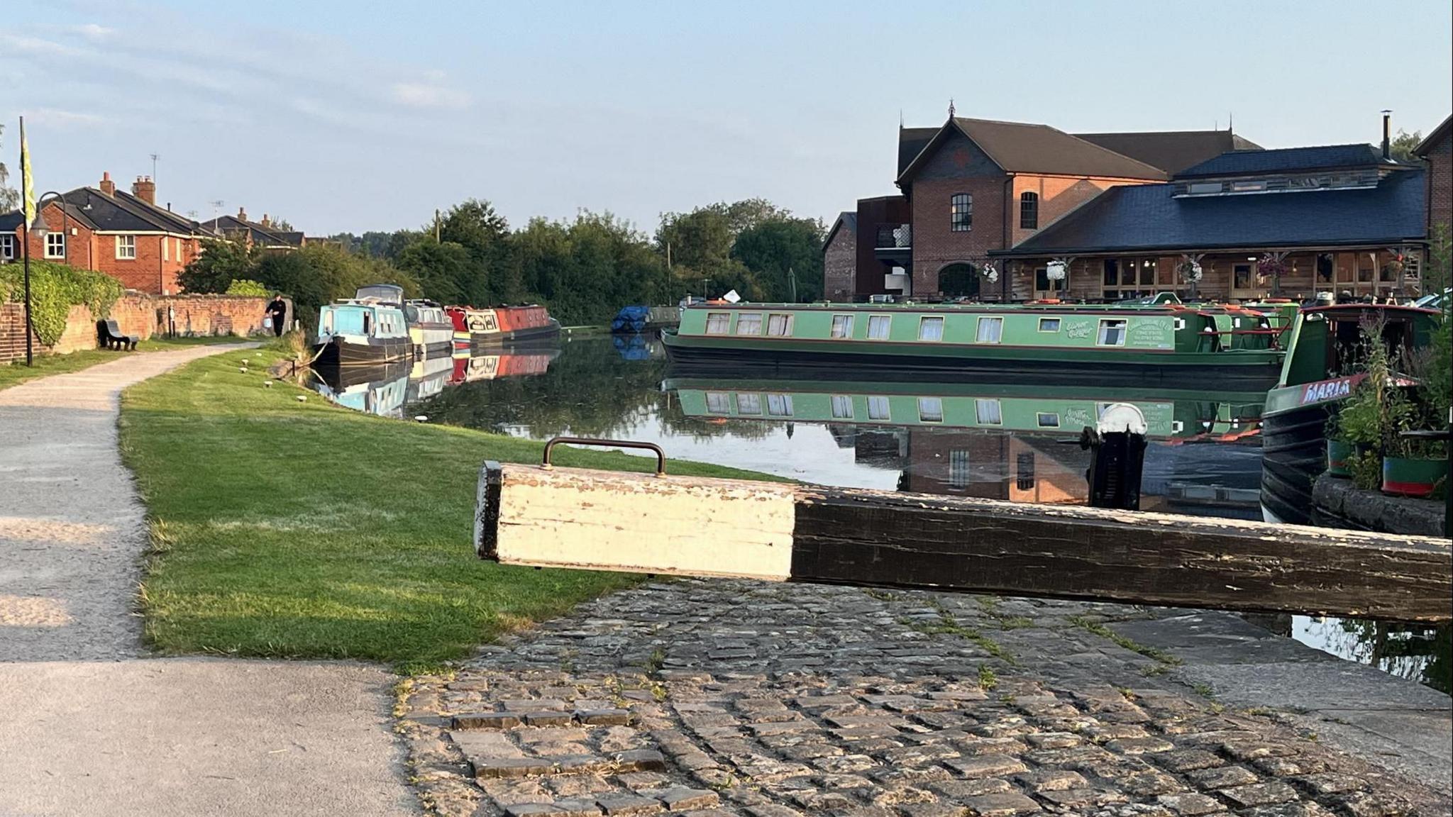 A lock gate in the foreground, with a number of boats moored in the pool and along the bank behind. In the distance is the Crown Wharf pub.