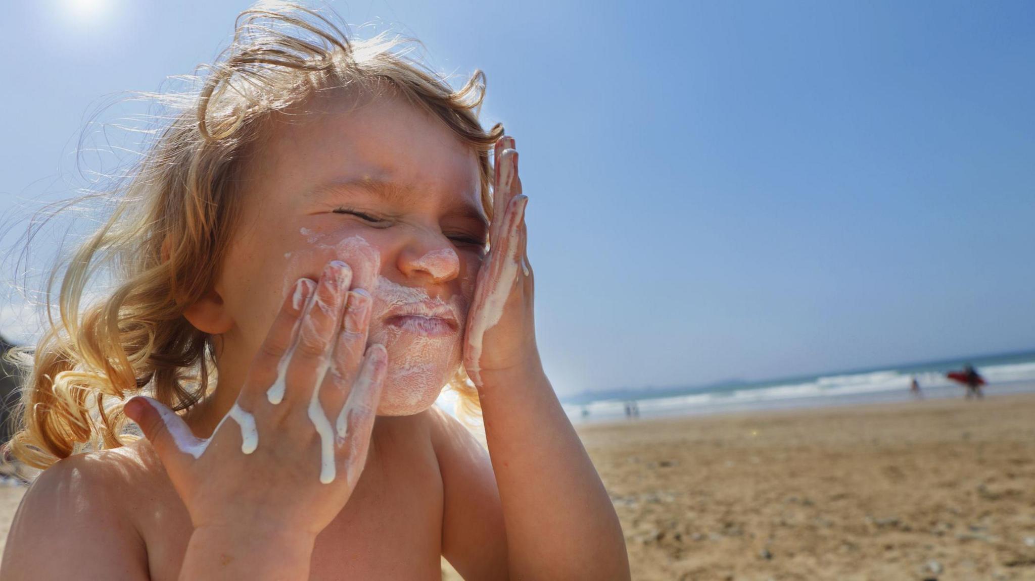 A blonde child with sunscreen dripping down her hands applies it to her face. A beach can be seen in the background.