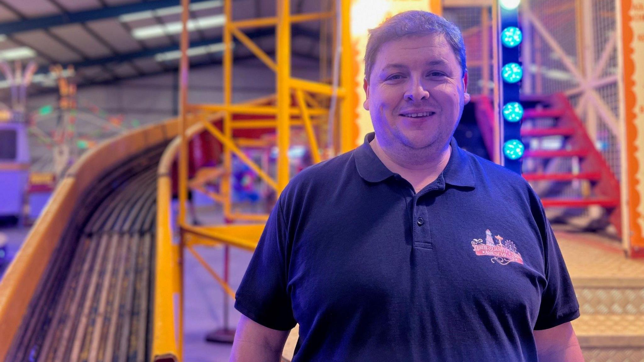 Leslie Danter is smiling at the camera and is standing in front of the yellow helter skelter which has bright blue lights on it.