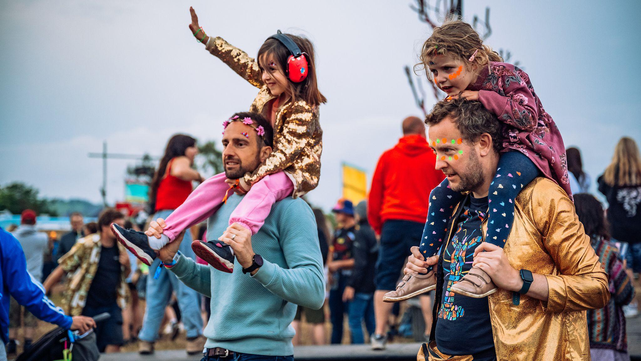 Two men in facepaint and a flower crown carrying children on their shoulders