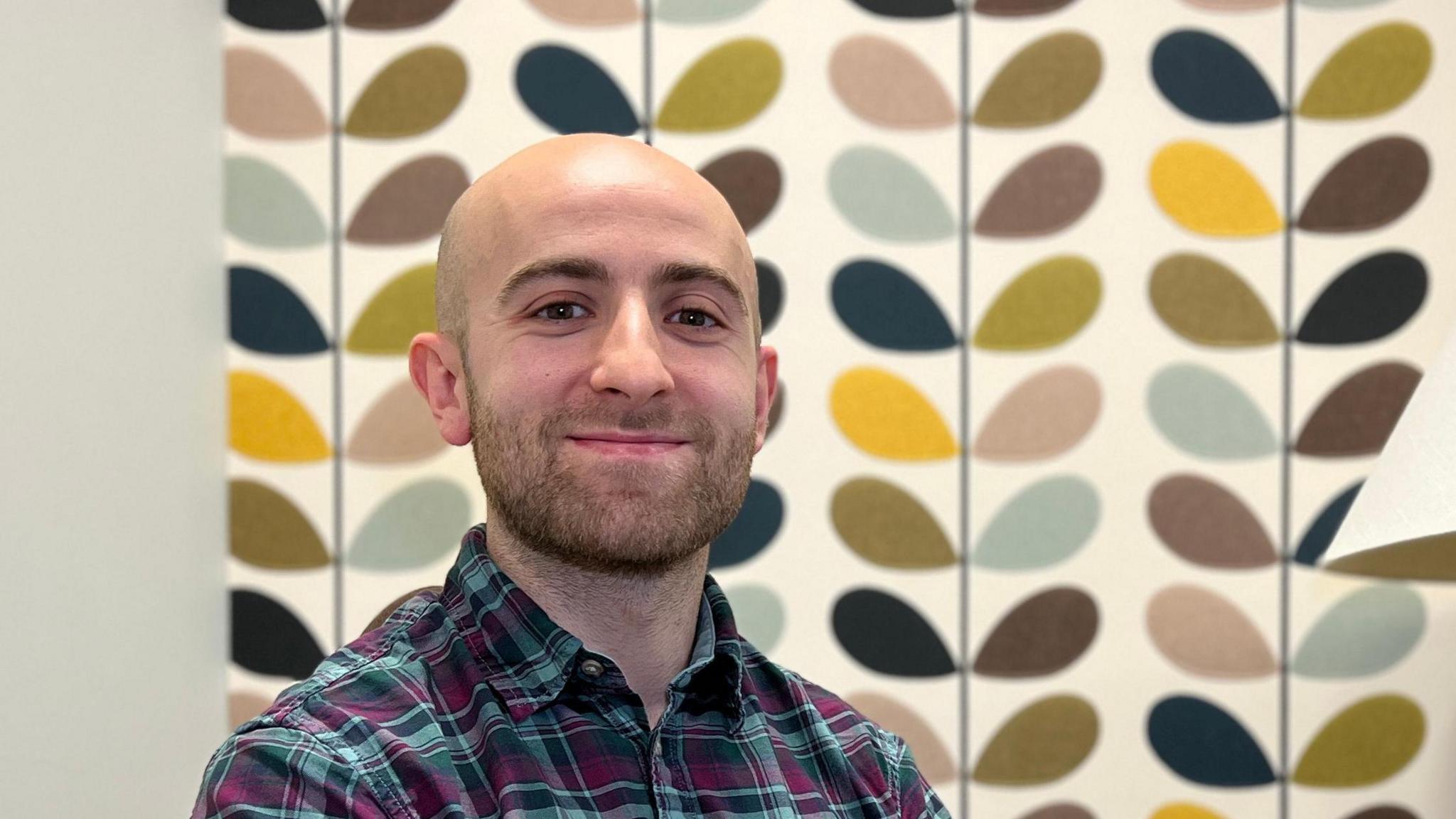 Jered looks at the camera and smiles. He's sitting on a green sofa chair and is wearing a pleated shirt and the sleeves are rolled up. There is a light to his right and the wall behind him has colour petals decorating the wall. Jered has a stubbly beard and is bald.