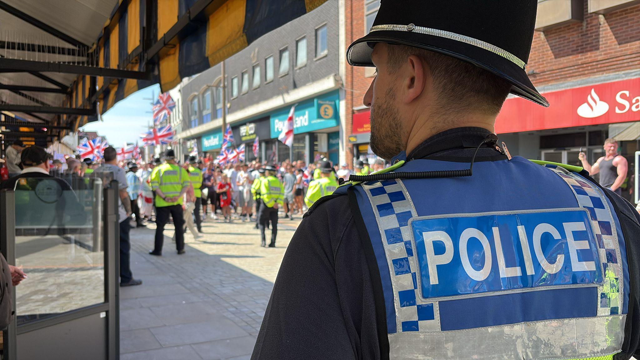 A police officer stands further back from the protest. He has a blue police vest on and a helmet. The protest further away is blurred in the photograph.